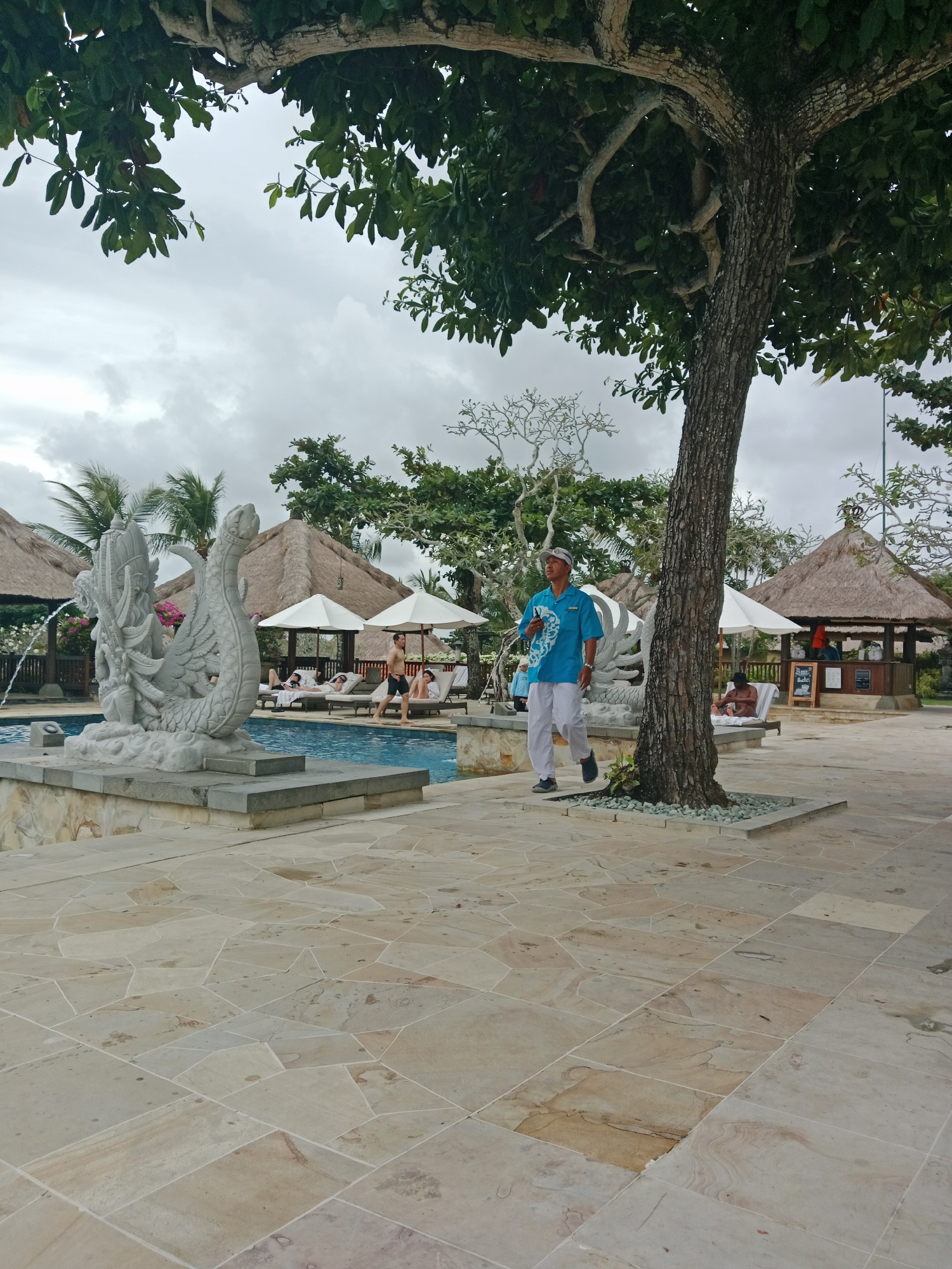 A man in a blue uniform and white pants walks across a paved patio in front of a swimming pool at a resort in Kuta Selatan, Indonesia. The man is wearing a white cap and appears to be holding a mobile phone in his left hand. To his left, a dragon-shaped fountain spouts water into the pool. In the background, thatched-roof cabanas and lounge chairs with white umbrellas are visible, some occupied by guests. Lush green trees and foliage surround the area, and the sky is overcast. The paved area is composed of irregular, tan-colored stone tiles.