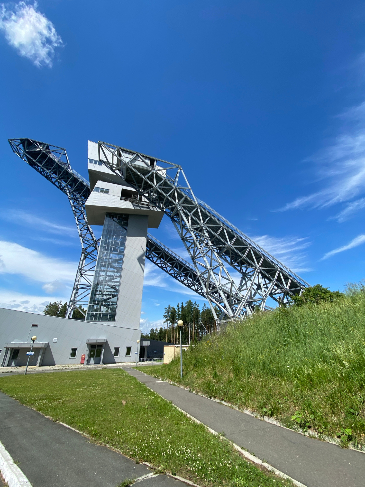 An outdoor, low-angle, full shot captures a modern ski jumping complex on a bright, sunny day in Prikhamsky, Russia. The complex features two massive, steel ramp structures that extend upwards and outwards against a vivid blue sky with wispy white clouds. A central grey tower with a glass elevator shaft connects the ramps. In the foreground, a paved walkway curves around a grassy area dotted with small white flowers. A grey building, part of the complex, is visible to the left, with several windows and two doors. To the right, a steep, grassy incline is covered in lush green grass and vegetation, with trees lining the background. The scene is devoid of people or any discernible activity.
