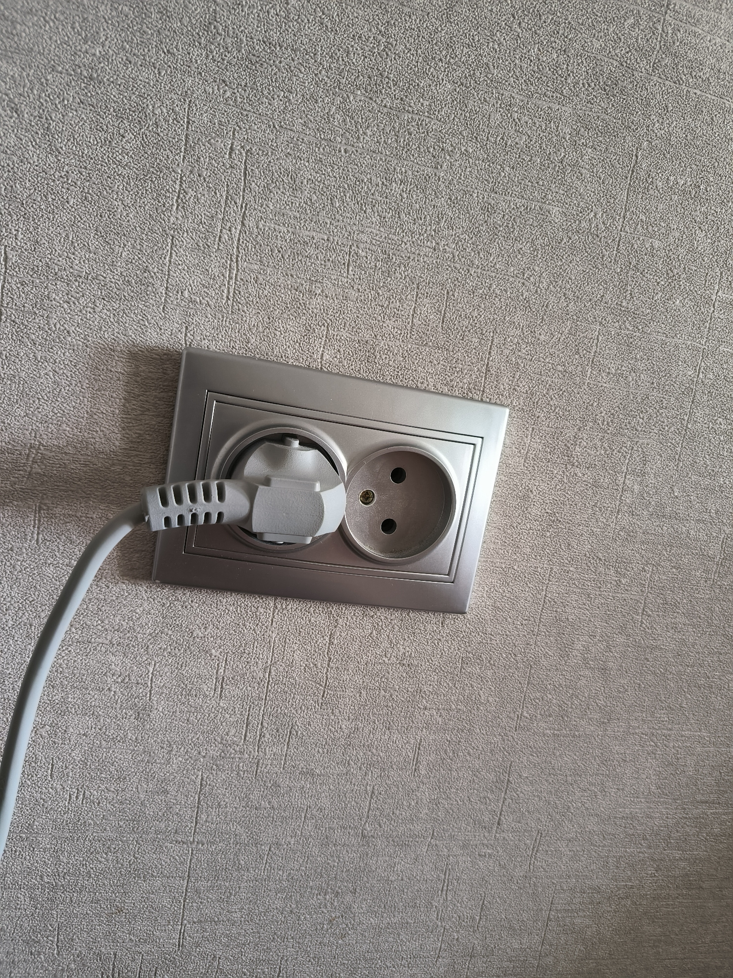 A power cord with a white plug is inserted into a dual electrical outlet mounted on a textured, light gray wall. The outlet itself has a silver metal frame. The wall has a repeating pattern of small bumps and subtle vertical lines, suggesting wallpaper or textured paint. The lighting appears to be artificial, casting a soft shadow to the left of the outlet. The image is taken from a slightly angled perspective, looking down at the outlet.