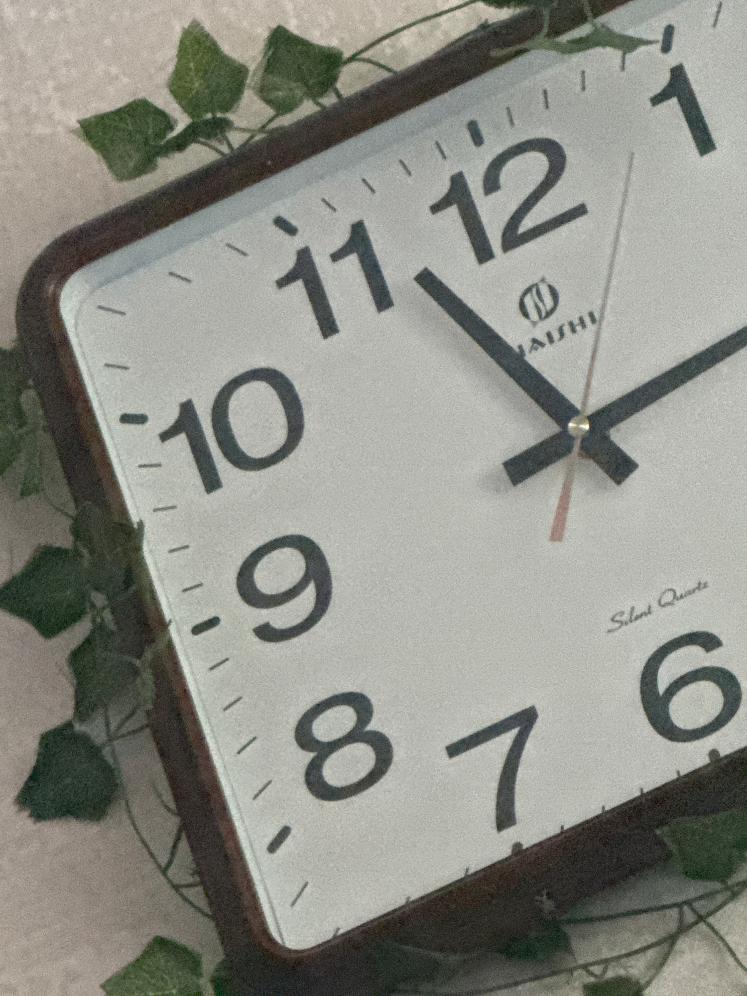 A square analog clock with a dark brown frame is mounted on a textured surface. The clock face is white with black Arabic numerals indicating the hours. The hour hand is pointing at approximately 4, and the minute hand is pointing at approximately 40. The second hand is pink and is pointing at approximately 39. The clock has the brand name "NAISHI" and "Silent Quartz" printed on its face. Green ivy vines are draped over the top and sides of the clock. The background is a light-colored, textured surface. There are no people or any discernible city elements.