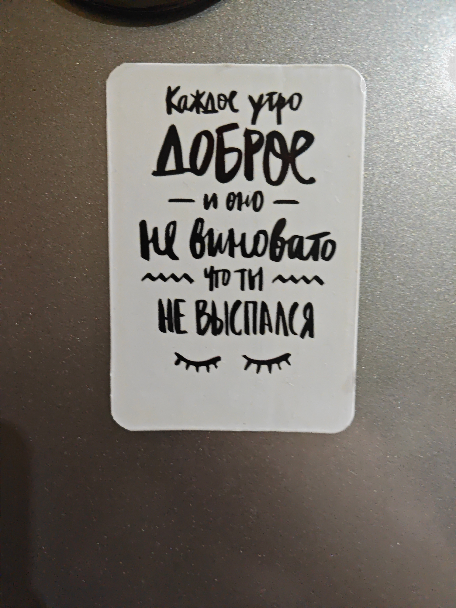 A white rectangular sticker with rounded corners is affixed to a metallic, textured surface, likely a refrigerator. The sticker contains hand-drawn text in black ink. The text reads: "Каждое утро ДОБРОЕ - и оно НЕ ВИНОВАТО ЧТО ТЫ НЕ ВЫСПАЛСЯ". Below the text, there are two stylized drawings of closed eyes with eyelashes. The metallic surface appears to be a neutral gray color and has a subtle sparkle effect. The lighting suggests it is an indoor setting. No people are visible in the image.