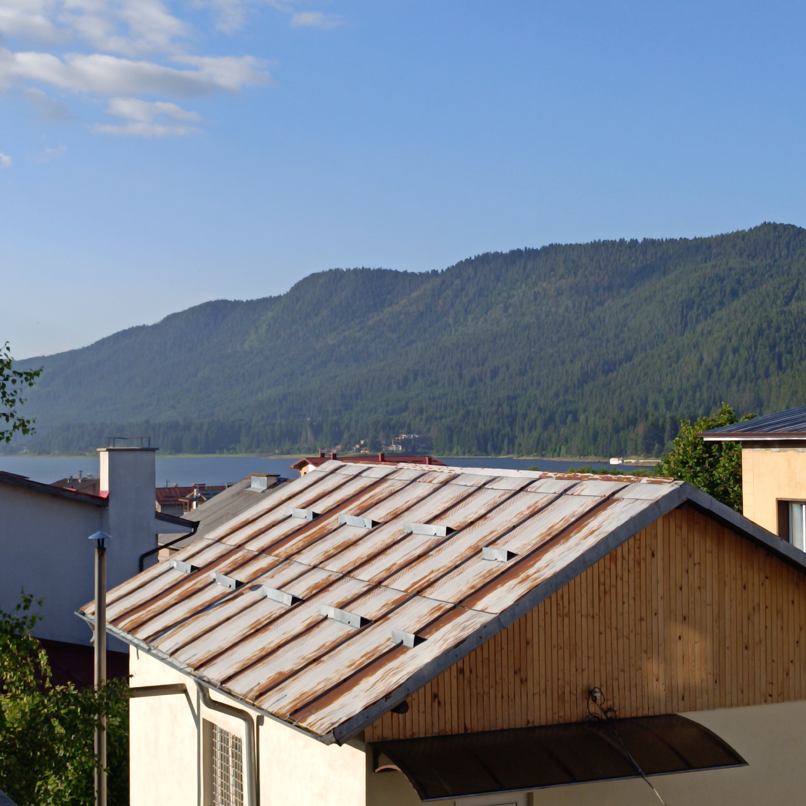 The image depicts a landscape view from a slightly elevated position in Sarnitsa, Bulgaria, showcasing a rural or semi-rural residential area with a backdrop of a forested mountain range and a body of water. In the foreground, a rustic metal roof of a building with visible rust and condensation runs diagonally across the frame. Adjacent to this structure, another building's cream-colored exterior wall is partially visible, featuring a window with a patterned grate and a tall, grey chimney. To the right, a section of a light yellow building with a dark-framed window is seen.
The middle ground is dominated by several rooftops, some with reddish tiles and others with grey metal. A prominent white chimney stands tall among these structures. Beyond the buildings, a calm blue body of water, likely a lake or reservoir, stretches across the image, reflecting the clear sky. In the distance, a majestic, heavily forested mountain range rises, its slopes covered in dense green trees. The sky is a bright, clear blue with a few wispy white clouds. The overall impression is one of a serene, sunlit day in a mountainous region of Bulgaria. There are no people or animals visible in the scene.