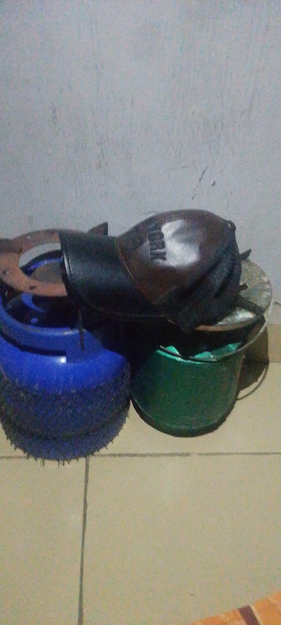 The image depicts a still life scene, likely indoors, featuring several objects stacked or placed together. In the foreground, there are two containers. The one on the left is a blue bucket, cylindrical in shape, with a textured outer surface that appears to be made of a rough material or pattern. To its right is a green bucket, also cylindrical, with a metallic rim.
Resting on top of these buckets is a brown object, possibly a shoe or a boot. It has a dark strap across it and a shiny surface with the letters "ORK" clearly visible, suggesting it might be a brand name. Underneath the brown object and partially visible, there appears to be some metallic or rusted material, possibly part of another object or a discarded component.
The background consists of a textured, light-colored wall, possibly concrete or plaster, with some faint marks or discoloration. The floor is a tiled surface, with visible grout lines forming a grid pattern. The lighting appears to be artificial, casting shadows that suggest the objects are illuminated from above and to the side. The overall impression is one of everyday objects, perhaps stored or left in a utility area. There are no people or discernible actions taking place in the image. The location in Jauro Yinu, Nigeria, is not directly evident from the visual information presented in the image.