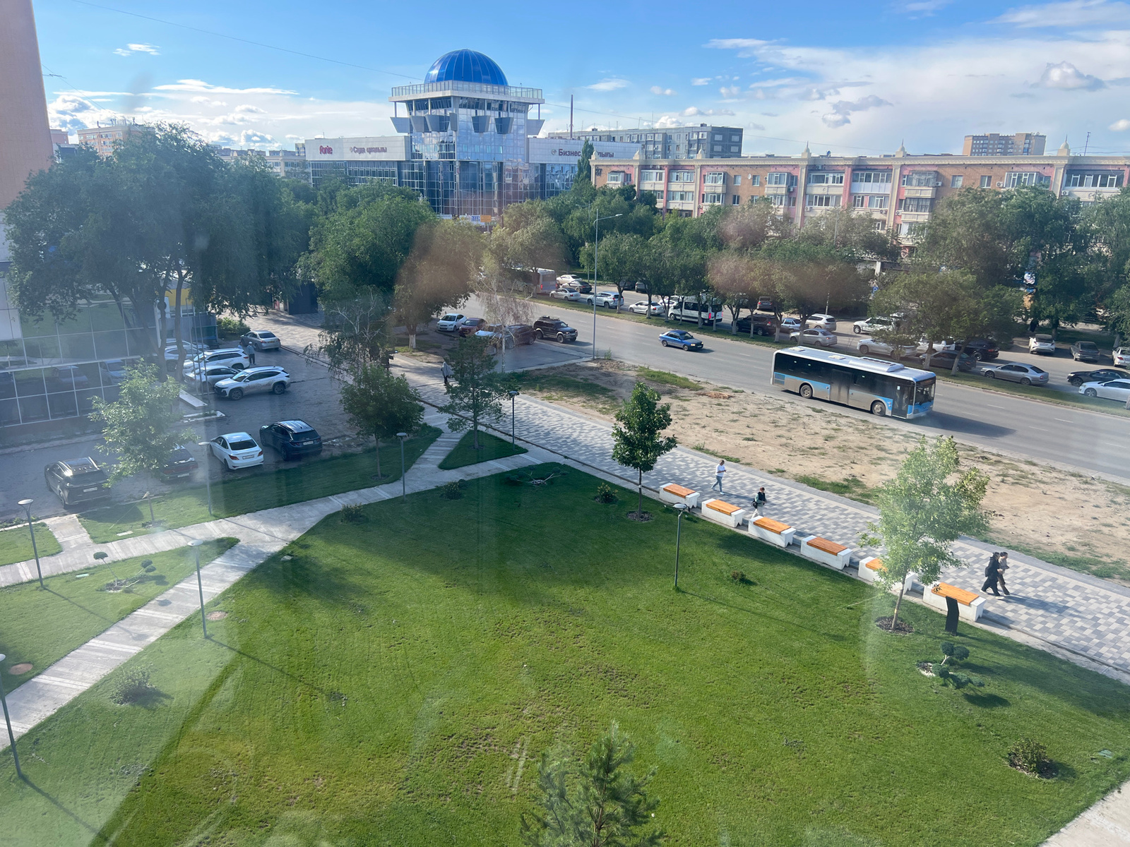 This is an aerial view of a street and a park in Aktobe, Kazakhstan. The scene depicts a sunny day with scattered clouds in a blue sky. On the left side of the image is a modern building with a blue dome, likely a public building or business center. Across the street, there are several multi-story residential buildings with balconies and various colored facades. A paved road runs horizontally across the middle of the image, with cars and a bus driving on it. On the right side of the road is a park area with green grass, trees, paved walkways, and modern benches. Several people are visible walking in the park and on the sidewalk. The overall scene suggests a well-maintained urban environment with both residential and commercial areas.