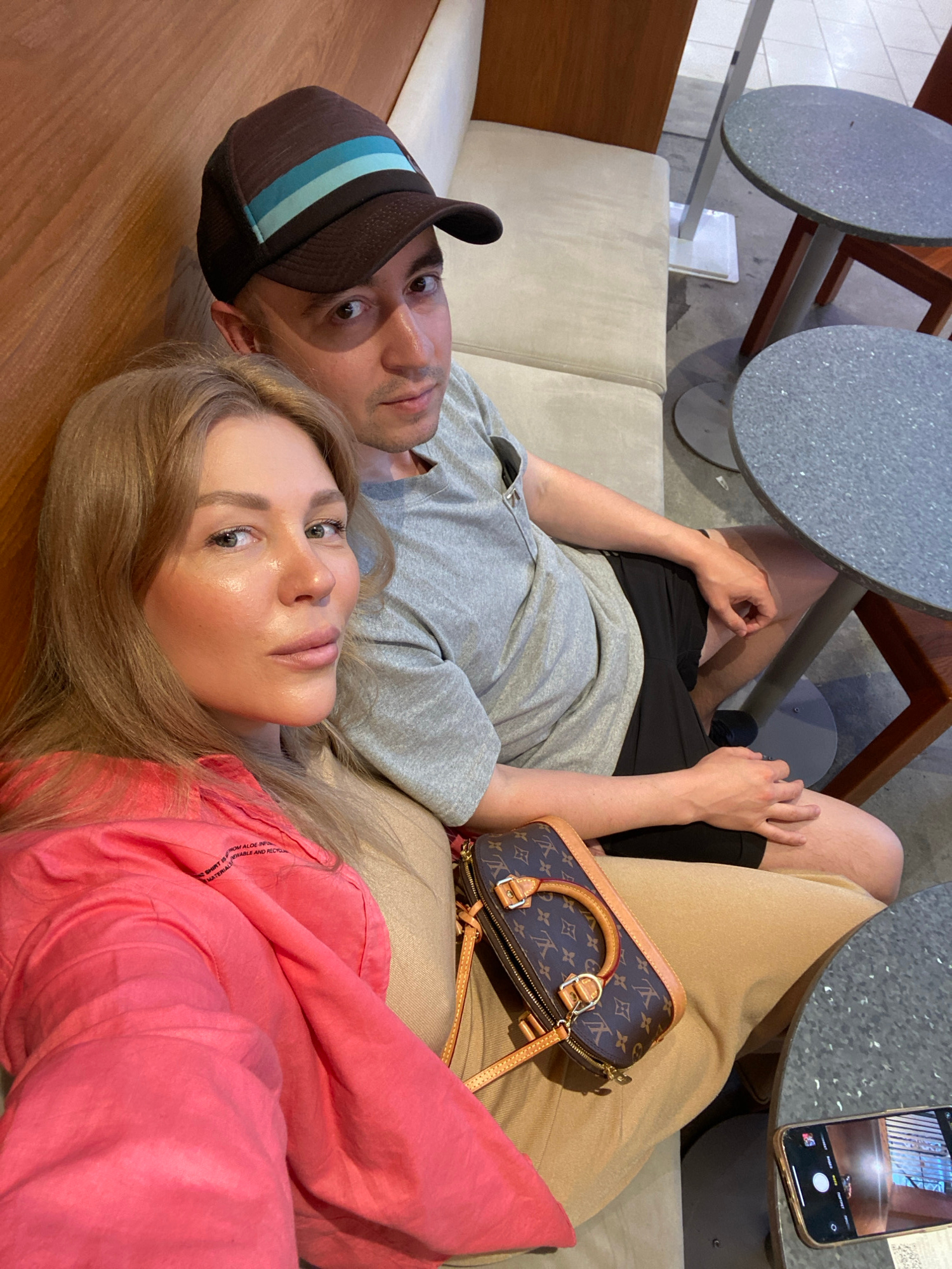 A medium shot captures a couple sitting on a light-colored sofa in what appears to be a cafe or restaurant. The woman, on the left, has blonde hair and is wearing a bright pink shirt. The man, to her right, is wearing a baseball cap and a grey t-shirt with black shorts. A Louis Vuitton handbag is placed on the woman's lap. In the background, there are round tables and chairs, suggesting an indoor seating area. The overall lighting suggests it is daytime. The image does not provide enough information to determine a specific location within Yekaterinburg, Russia.
