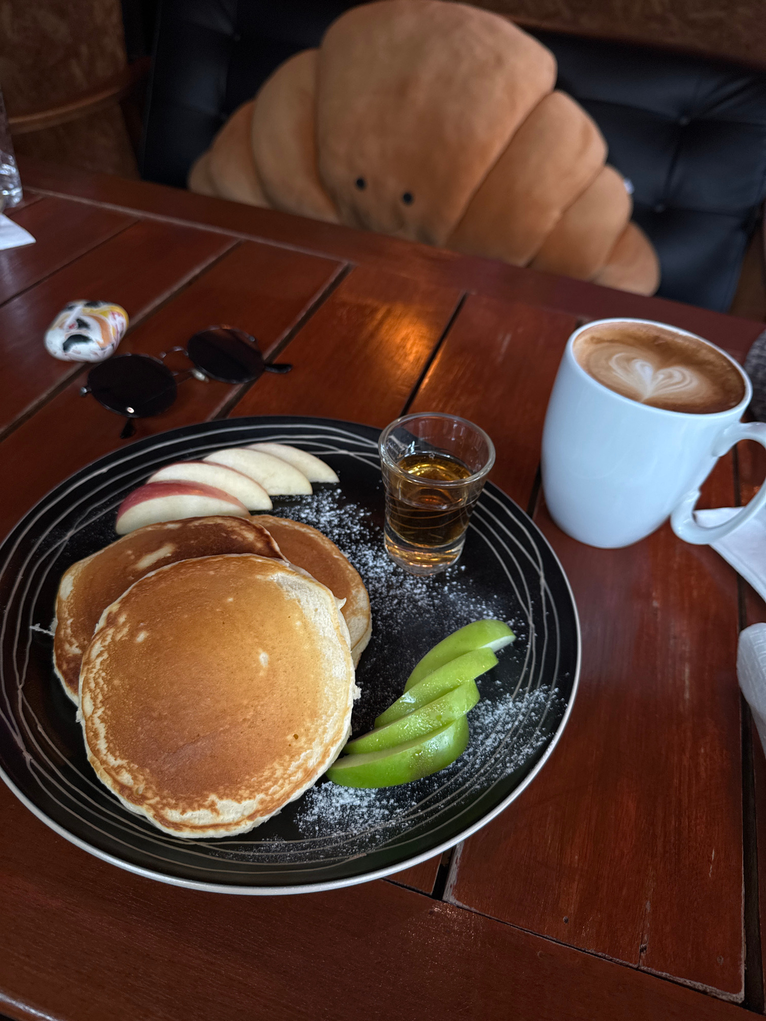 A breakfast scene is depicted on a wooden table in Rawai, Thailand. In the foreground, a black plate holds three golden-brown pancakes dusted with powdered sugar, accompanied by sliced red apples and bright green apple slices. A small shot glass filled with amber liquid, likely syrup, sits next to the pancakes. To the right, a white mug with latte art in the shape of a heart is filled with coffee. In the background, a plush croissant-shaped pillow rests on a black chair, and on the table, a pair of dark sunglasses and a small, colorful decorative stone are visible. The lighting suggests an indoor setting, possibly a cafe.