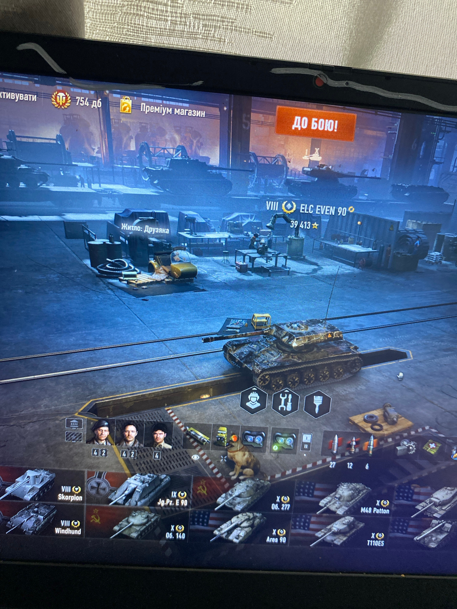 The image shows a computer screen displaying a video game, specifically a tank simulation game. The game interface shows a garage or hangar area with various tanks. In the foreground, a detailed model of a tank is displayed on a ramp. Below this, a row of icons and smaller tank images represent selectable vehicles. The game's menu bar at the top indicates options like "Activate," "Premium Shop," and a "Battle!" button. The background depicts an industrial-like setting within the game. There is no indication that this scene is located in the city of Szczecin, Poland. The image is a photograph of a computer monitor, with some glare and reflections visible on the screen.