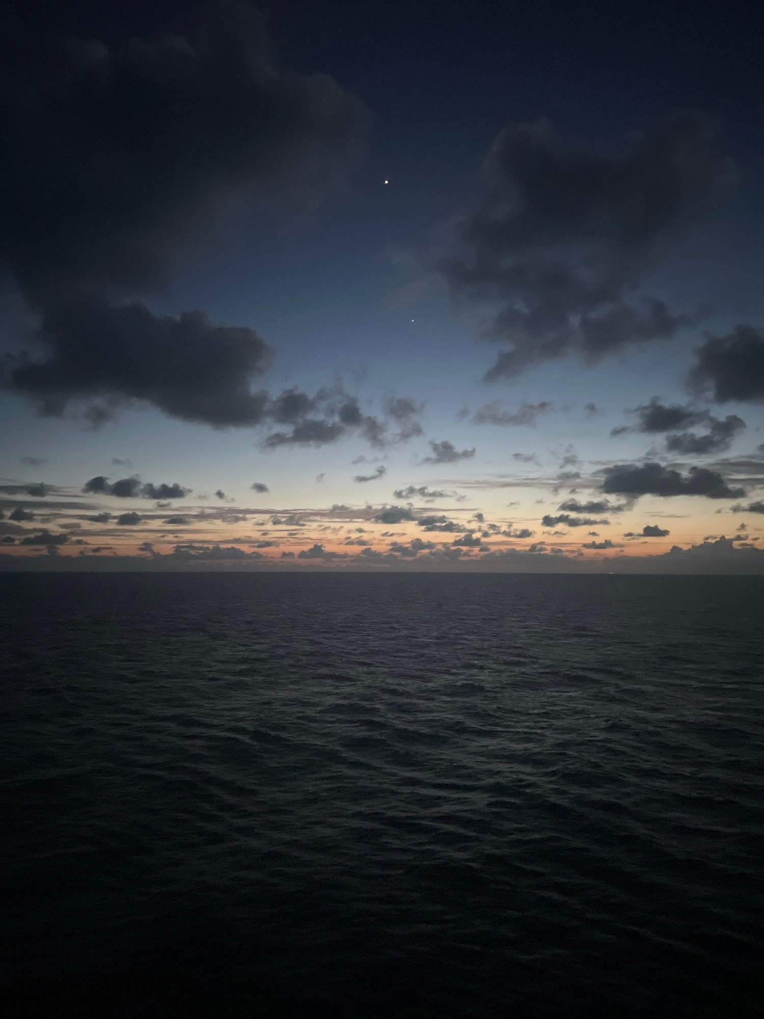 A serene ocean sunset unfolds under a dramatic sky, casting a somber yet beautiful light over the scene. The sky transitions from a deep, inky blue at the top to soft hues of orange and pink near the horizon, with scattered clouds adding texture and depth. Two celestial bodies, likely planets or bright stars, are visible in the upper part of the sky. The ocean below is a dark, rippling expanse, reflecting the muted colors of the sunset. The image evokes a sense of vastness and tranquility, capturing a moment of transition between day and night at sea.