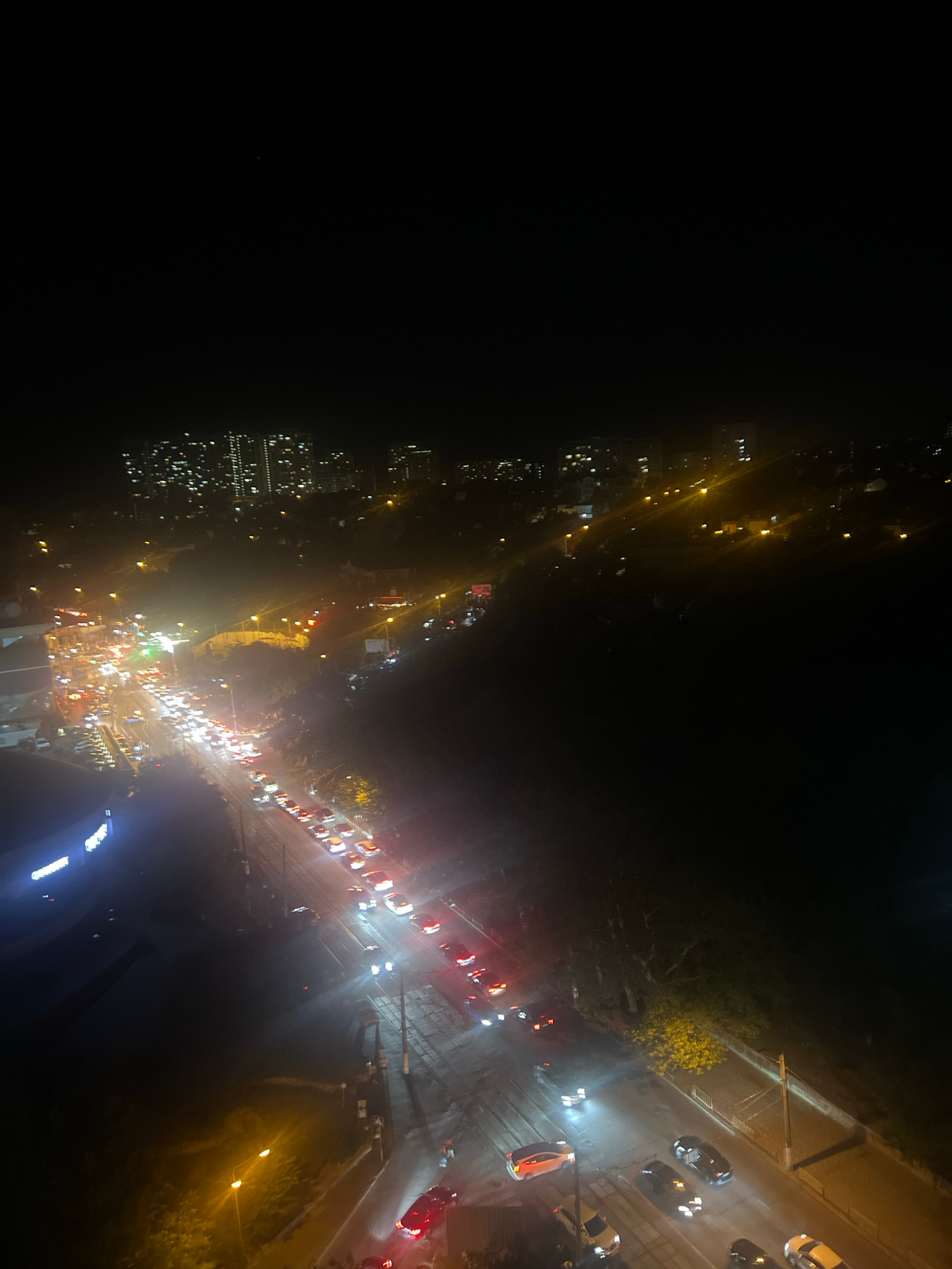 An elevated, nighttime, wide-angle shot captures a congested urban road in Odesa, Ukraine. The dark sky is illuminated by a dense array of city lights, including numerous apartment buildings in the background and streetlights lining the road. The road itself is filled with a continuous stream of vehicles, their headlights and taillights creating streaks of light due to the long exposure. The traffic appears to be moving slowly, indicating a traffic jam. Trees and foliage line the sides of the road, their details obscured by the darkness and ambient light. A few street lamps cast a yellowish glow on the scene. The image is characterized by a high level of visual noise and a slight blur, likely due to the low light conditions.