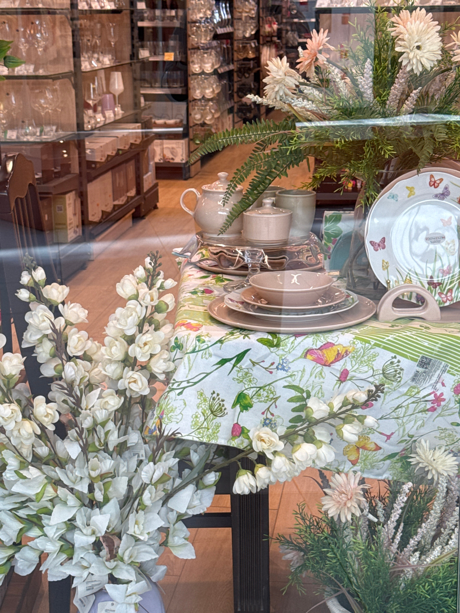 The image displays a store window featuring a table set with white and pink floral arrangements. A tablecloth with a butterfly and floral pattern is laid out, on which a teapot, sugar bowl, and stacked plates with bowls are arranged. A decorative plate with butterflies is positioned to the right of the table setting. Behind the table, shelves are visible stocked with glassware and other retail items. The scene is brightly lit, suggesting an indoor retail environment. There are no people or animals present in the image.