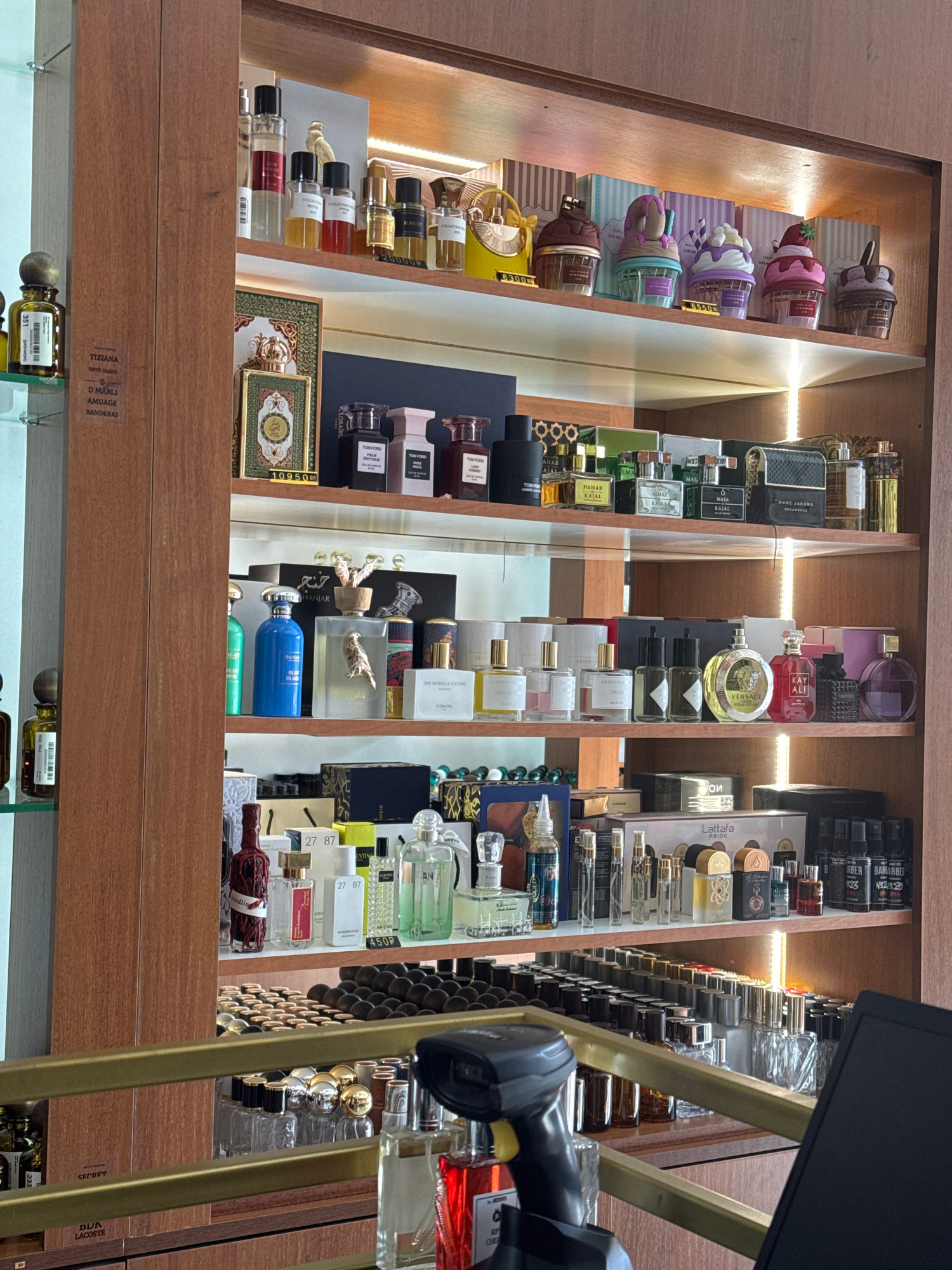 This image shows a retail store interior, specifically a display shelf filled with various perfume bottles. The shelves are made of wood, and there are interior lights illuminating the products. The perfumes are arranged in rows and on different levels, with a variety of shapes, sizes, and colors of bottles. Some bottles are in their original packaging, while others are displayed individually.
A sign on the left side of the image, on the wooden frame, contains text in English, indicating product names and potentially brands. Some of the text is partially obscured but includes recognizable words like "TIZIANA," "D MARLI," "AMUAGE," and "BANDERA."
The lower part of the image shows more shelves with perfumes and what appears to be a checkout counter with a barcode scanner and a black monitor. The overall impression is of a well-stocked perfume shop.
While the image depicts a retail environment with numerous perfume products, there is no information within the visual content of the image to determine its specific location within the city of Krasnodar, Russia. The text on the bottles and shelves are in English, but this does not confirm or deny the location.