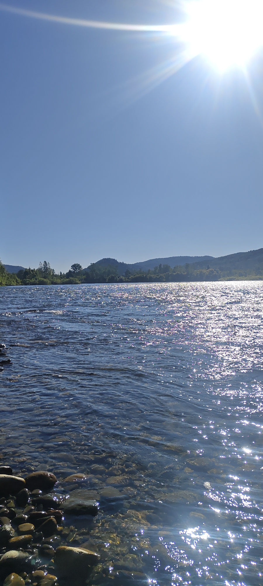 The image depicts a wide, clear river flowing through a natural landscape under a bright, sunny sky. The sun, positioned in the upper right corner, casts brilliant reflections on the water's surface, creating a shimmering effect. The riverbed is visible in the foreground, composed of smooth, rounded stones. Along the far bank, a lush green treeline and rolling hills form the horizon, under a cloudless blue sky. The location is identified as Mezhdurechensk, Russia.