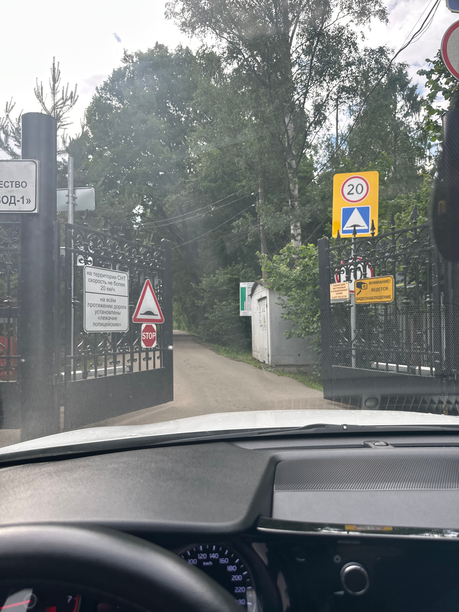 The image shows a view from inside a car, looking forward through an open black metal gate. The gate is ornate and appears to be the entrance to a property. On the left side of the gate, there is a black cylindrical post with a white sign that reads "ЕСТВО / ЗОД-1»". Further into the property, a narrow paved road leads into a wooded area.
Attached to the gate itself are several signs. On the left leaf of the gate, there is a white rectangular sign with black text that states: "на территории СНТ / скорость не более / 20 км/ч" (on the territory of SNT / speed not more than / 20 km/h) and "на всем / протяжении дороги / установлены / «лежачие / полицейские»" (along the entire / length of the road / are installed / "speed bumps"). To the right of this sign, there is a triangular road sign with a red border and a black depiction of a speed bump, indicating a hazard. Below this hazard sign is a circular red and white "STOP" sign.
On the right leaf of the gate, there is a yellow rectangular sign with black text and a camera icon, reading "ВНИМАНИЕ! / ВЕДЕТСЯ / ВИДЕОНАБЛЮДЕНИЕ" (ATTENTION! / VIDEO SURVEILLANCE IS BEING / CONDUCTED). To the left of this sign, there is another smaller yellow sign with black text and a phone number. Above these signs, a circular yellow traffic sign displays the number "20" inside a red circle, indicating a speed limit of 20 km/h. Below this, there is a blue triangular sign with a white arrow pointing upwards, which likely indicates an upcoming obstacle or change in the road.
In the background, beyond the gate, there are trees and a small white building with a garage door. The sky is overcast. The car's dashboard and steering wheel are visible in the foreground. The speedometer shows a speed of approximately 0 km/h. The image appears to have been taken in Krasnogorsk, Russia.