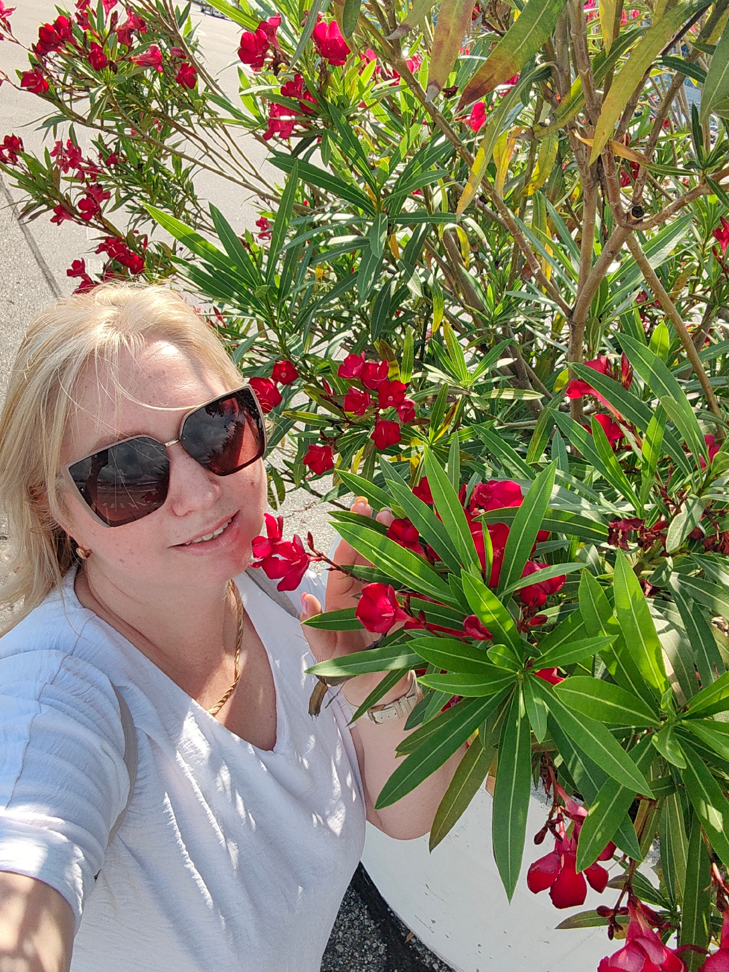 A woman with blonde hair and sunglasses is standing next to a flowering bush. She is wearing a white shirt and a gold necklace. The bush has red flowers and green leaves. The background is blurred and appears to be an outdoor setting. There are no indications of the specific location within the city of Sirius, Russia, in the provided image.
