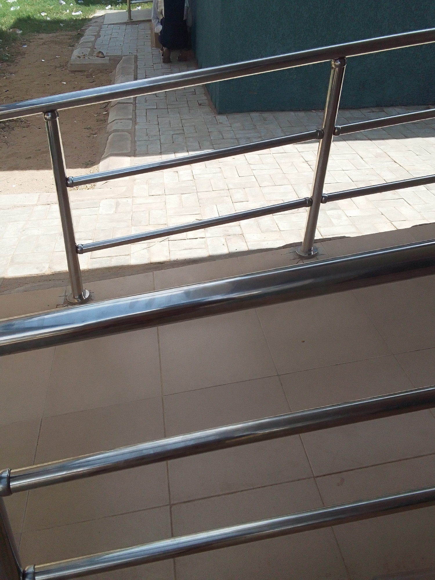 This media file shows a close-up view of a metal handrail on a ramp or walkway. The handrail is made of polished stainless steel, with a thicker top rail and a thinner bottom rail. It is supported by vertical posts that are also made of stainless steel. The handrail is angled, suggesting it is part of a ramp designed for accessibility.
In the background, there is a paved walkway made of rectangular stone or concrete tiles. To the left of the walkway, there is an area of dirt and dry grass, with some litter visible. To the right, there is a dark green wall.
Further in the background, a glimpse of two people can be seen. One person appears to be wearing dark clothing and brown shoes. The other person's attire is less clear. They are standing on the paved walkway.
The lighting suggests it is daytime, with shadows cast on the walkway, indicating sunlight. The overall scene appears to be outdoors, possibly in a public area like a park, school, or commercial building entrance. There is no visible text in the media file.