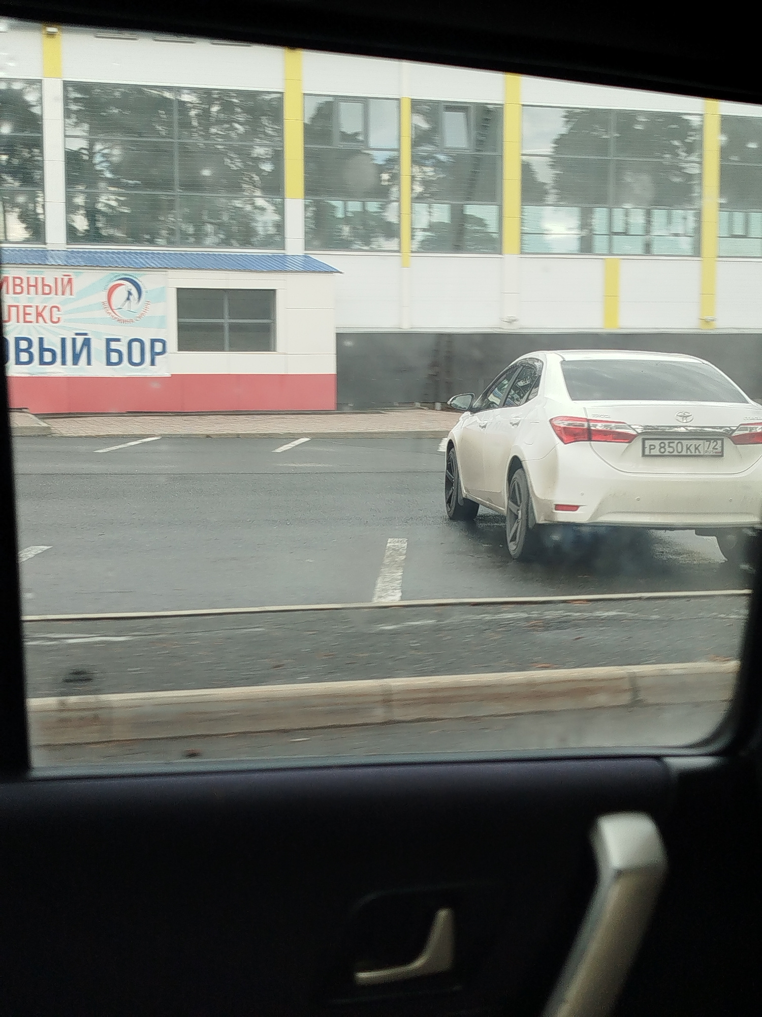The image is taken from inside a vehicle, looking out through the window. In the background, a modern white building with large windows is visible, with yellow vertical accents. A banner with Russian text and a circular logo is displayed on the side of the building. To the right of the building, a white sedan is parked on an asphalt surface marked with white lines, suggesting a parking lot or road. The license plate of the car is partially visible and reads "P 850KK 72". The scene appears to be in an urban or suburban area.