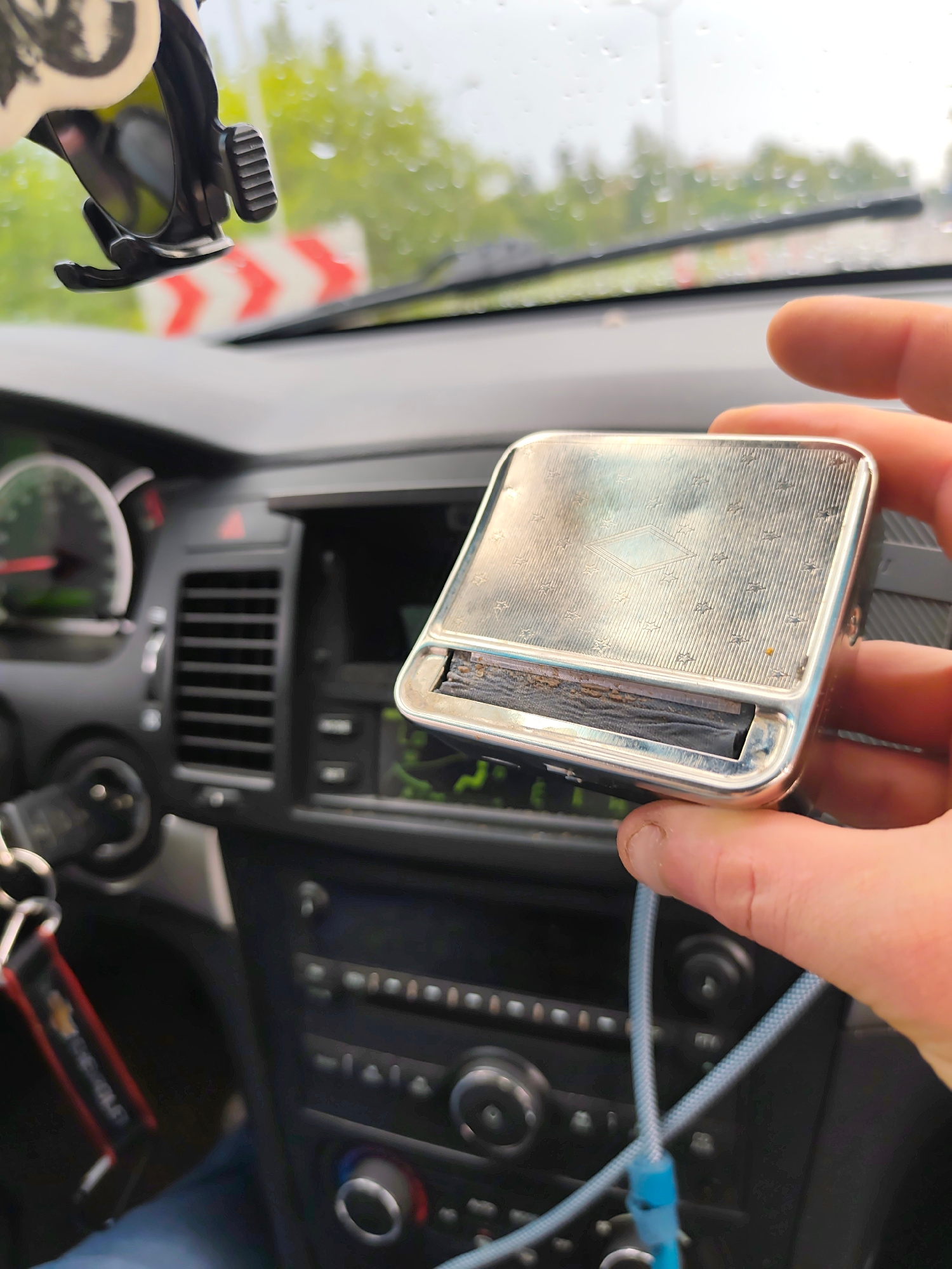 A close-up shot shows a hand holding a silver cigarette rolling machine inside a car. The car's dashboard is visible, with a stereo system, air vents, and part of the steering wheel. The rolling machine has an intricately engraved lid with a diamond pattern and small star-like imprints. The rolling mechanism itself appears worn and contains dark material, likely tobacco. A light blue cord, possibly a charger or auxiliary cable, is attached to the rolling machine.
In the background, through the car's windshield, a blurry view of the outdoors reveals a cloudy day with visible raindrops on the glass, suggesting it might be raining or has recently rained. A chevron-style road sign with red and white stripes is also partially visible outside the car, indicating a roadway.
The scene is set within a car, likely in an urban or suburban environment given the road sign. The overall mood is casual and ordinary, capturing a moment of use or inspection of the rolling machine. There is no visible text on the rolling machine itself, but a red and black keychain with a "CHEVROLET" logo is attached to a set of keys on the left side of the dashboard, providing a brand cue for the car. The image was taken in Szczecin, Poland.