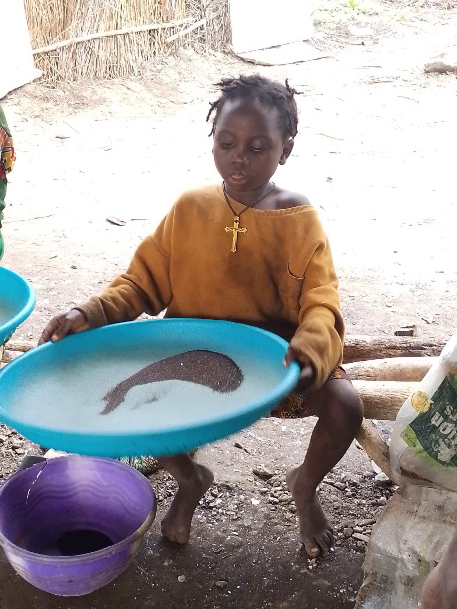 The media file shows a young child, likely a girl, of African descent sitting outdoors in what appears to be a rural setting in Otulu, Nigeria. She has dark skin and dark, short, braided hair. The child is wearing an oversized, mustard-yellow, long-sleeved sweatshirt that is off one shoulder, and a colorful patterned skirt. Around her neck, she wears a gold-colored cross pendant on a dark cord.
In her hands, the child holds a large, bright blue, shallow bowl filled with water and what appears to be fine, dark-colored sand or soil, with some lighter sand or material mixed in. She is holding the bowl in a way that suggests she might be sifting or washing the contents. To her left, another blue bowl, partially visible, and to her right, a large purple bowl or basin containing some dark liquid or material is on the ground. The ground itself is rough and appears to be dirt, gravel, and possibly some wet patches.
In the background, there is a rustic structure constructed from what looks like dried reeds or straw, supported by wooden poles. There are also some logs or branches placed horizontally behind the child. To the right of the child, a white and green bag with some visible text is leaning against a surface. The visible text on the bag includes "tion rs" and "TM 2014".
The overall scene suggests a moment of everyday activity, possibly involving the processing of materials from the earth. The lighting indicates it is daytime, likely with overcast or diffused sunlight given the lack of harsh shadows. The child's expression is one of focus on the task at hand, with a neutral or slightly serious demeanor. The setting and activity hint at a possibly resource-intensive or labor-intensive environment.