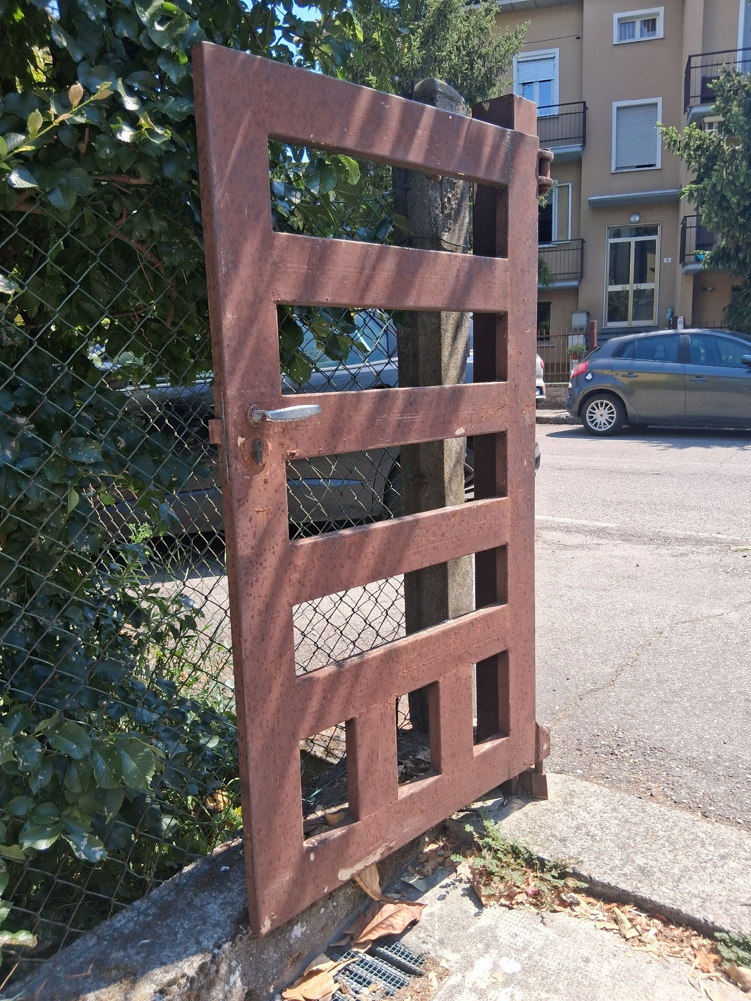 An opened, rusty brown metal gate, with a rectangular frame and horizontal bars, stands upright on a concrete sidewalk. The gate is attached to a concrete post and has a silver metal handle and a lock mechanism. Behind the gate, a chain-link fence is visible, with dense green foliage growing through it. A paved street runs alongside the sidewalk, with a parked grey hatchback car on the right side of the image. In the background, a multi-story building with balconies and windows is visible, set against a clear blue sky. The scene appears to be located in Colorno, Italy, on a sunny day.