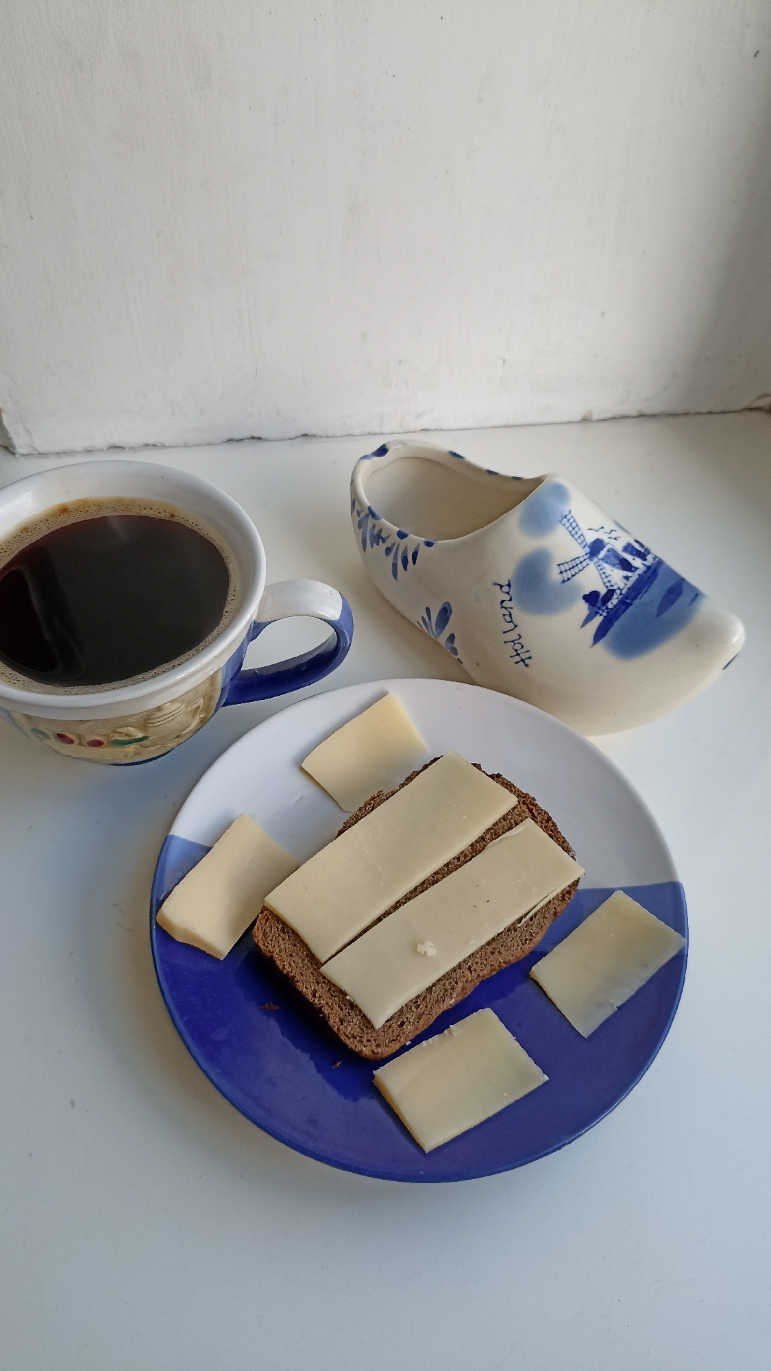 A close-up, top-down shot shows a rustic breakfast spread on a white surface, likely a windowsill or table, in Kapyĺ, Belarus. In the foreground, a blue and white plate holds a slice of dark bread topped with several slices of light-colored cheese. Three additional cheese slices are scattered around the bread on the plate. To the left of the plate, a white mug with blue trim is filled with dark coffee. Behind the plate and coffee cup, a decorative white ceramic clog with blue Dutch-style windmill imagery and the word "Holland" is visible. The background consists of a textured white wall.