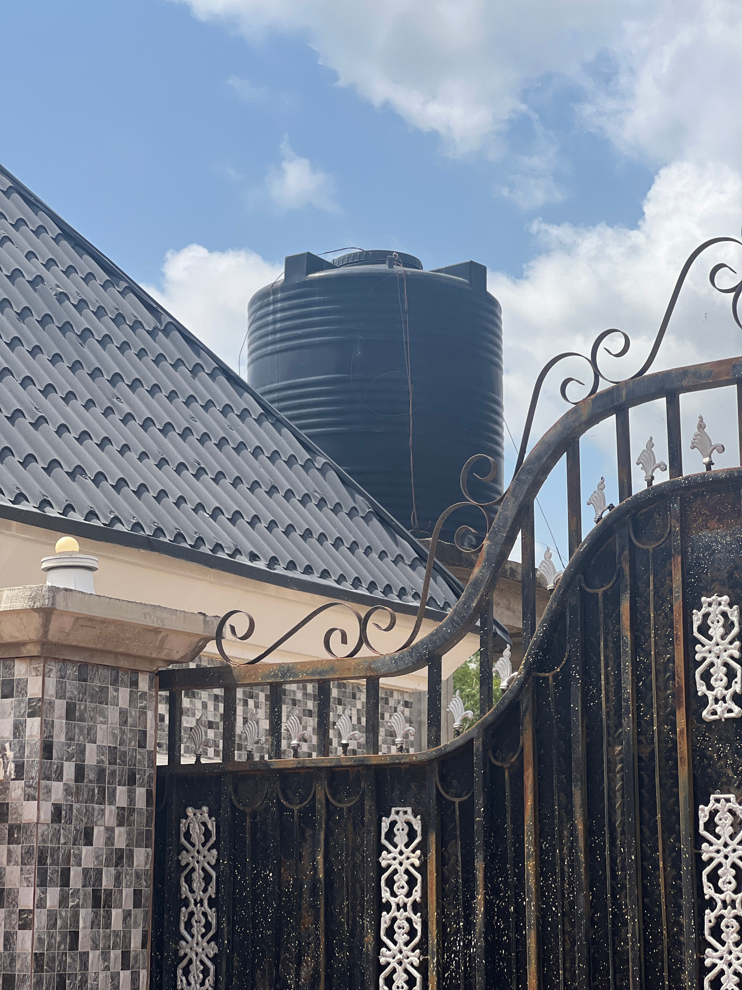 The image displays a low-angle view of a black water tank situated on the roof of a building, which has a dark, patterned roof. The building is adorned with patterned tiles and appears to be partly covered by an ornate black metal gate. The gate features decorative scrollwork and white filigree details. In the background, a bright blue sky with scattered white clouds suggests it is daytime with fair weather. The overall scene is of an exterior view of a building, possibly in a residential area in Howayi, Nigeria. No people or specific activities are depicted, and there is no visible text in the image. The lighting indicates it is daytime, likely sunny, given the brightness of the sky and the clear visibility of the objects.