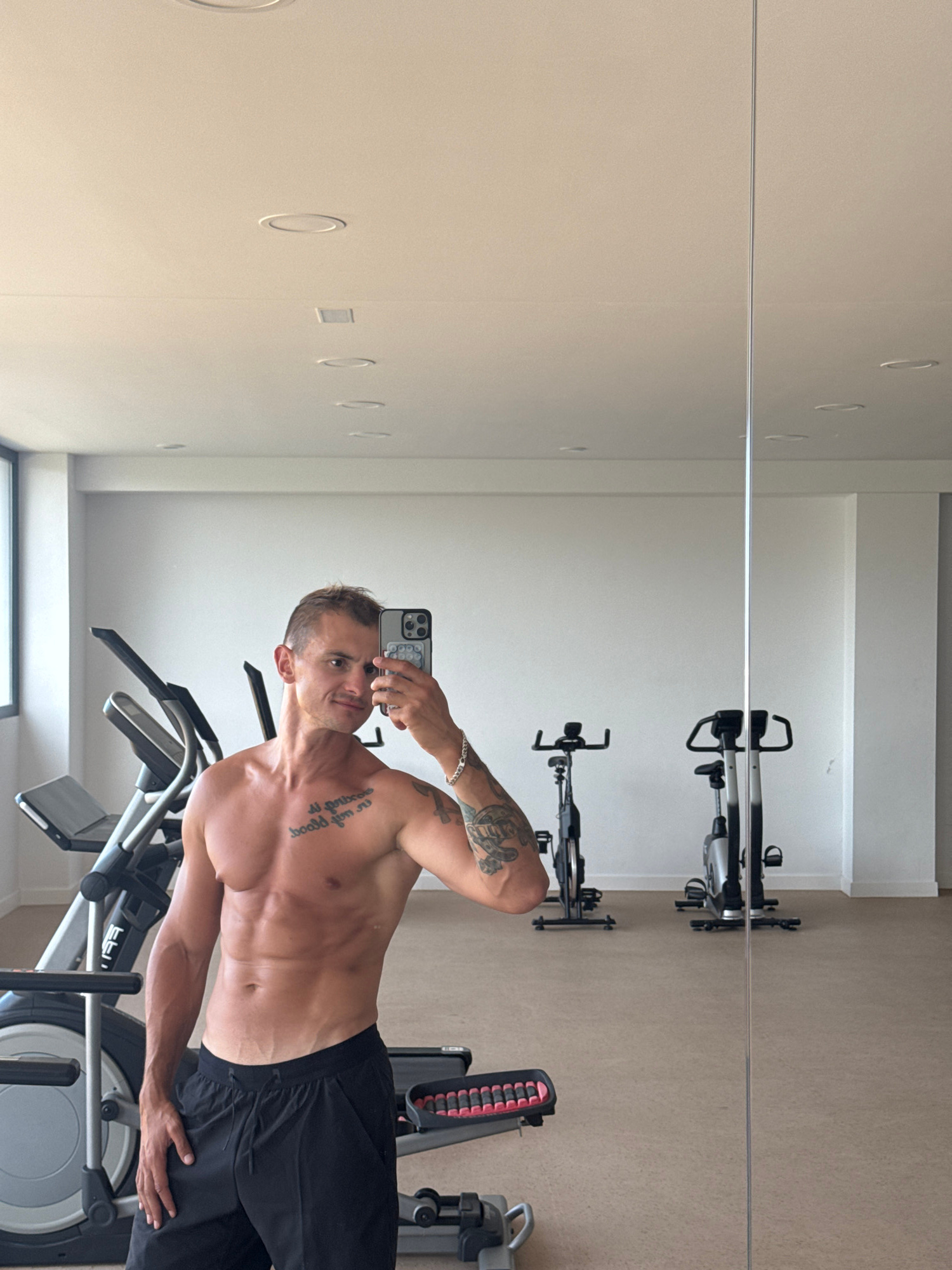 The image depicts a shirtless man with visible abs and tattoos posing for a selfie in a gym. He is wearing black athletic shorts. In the background, there are several pieces of exercise equipment, including an elliptical machine and two stationary bicycles. The gym appears to be well-lit, with recessed lighting on the ceiling and large windows. The floor is a light-colored carpet. The man is looking at his phone, which is held up to take the picture. The setting is consistent with a gym located in Finestrat, Spain, given the clean, modern aesthetic and the type of exercise equipment typically found in such establishments.