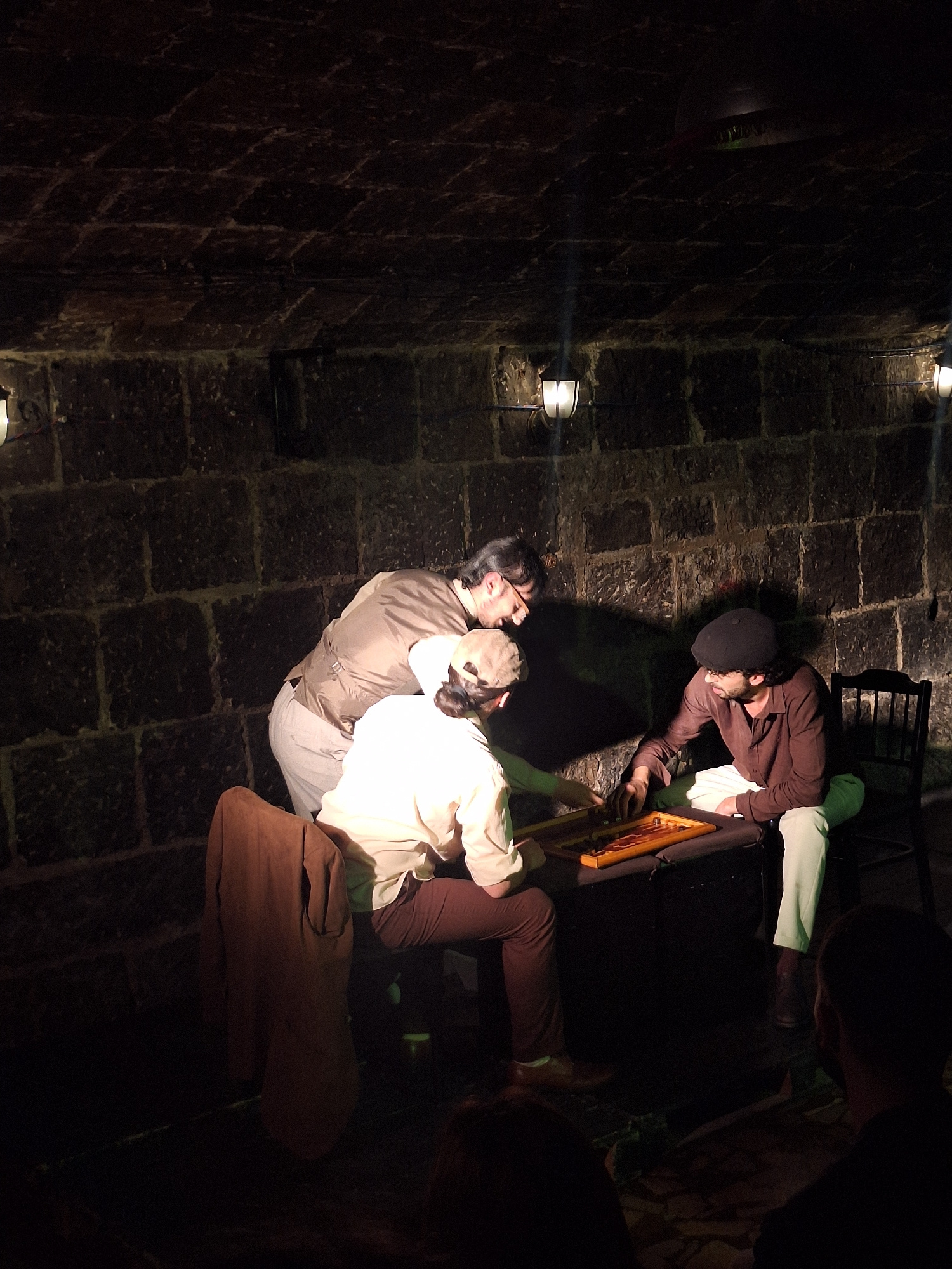 This media file captures a scene with several individuals engaged in an activity within a dimly lit, stone-walled setting that appears to be outdoors, possibly a courtyard or alleyway. The stone paving and rough-hewn walls suggest a historic or rustic location, consistent with the context of Gyumri, Armenia.
The primary subjects are at least four people, their faces partially obscured by the lighting and angles. Two individuals are seated at a low table, seemingly engrossed in a game or a shared task. One person, wearing a brown shirt and a dark cap, leans over the table, his hands active. Another person, dressed in lighter clothing, is also positioned at the table, with their head close to the other individual. A third person, in a light-colored top, is partly visible behind them. A fourth person's legs and torso are visible at the bottom right of the frame, covered by what seems to be a brown garment.
The overall mood is intimate and focused, with the dim lighting casting long shadows and highlighting the textures of the stone. The activity appears to be a game involving a wooden board with circular elements, perhaps a traditional game. The time of day is difficult to ascertain definitively due to the artificial lighting, but the dark surroundings suggest it is either evening or night. No text is visible in the image. The scene evokes a sense of quiet concentration and shared engagement in a historical setting.