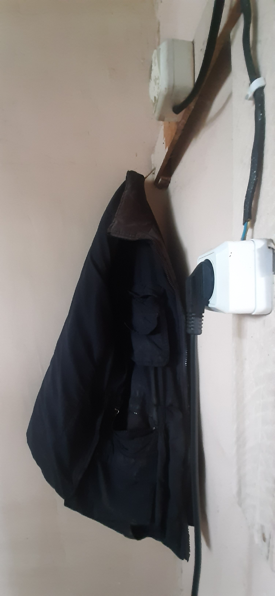 A black jacket hangs from a wooden peg on a light-colored wall. Two electrical outlets are visible on the wall. One outlet, located above and to the left of the jacket, appears to have a black cable plugged into it, with a portion of the cable secured by a white clip. The second outlet, located to the right of the jacket, has a black plug inserted into it. A thick black cable extends from this plug downwards. The wall itself appears somewhat worn and textured. The image is taken from a slightly low angle, looking up towards the hanging jacket and outlets. The scene is indoors. The location is Yessentukskaya, Russia.