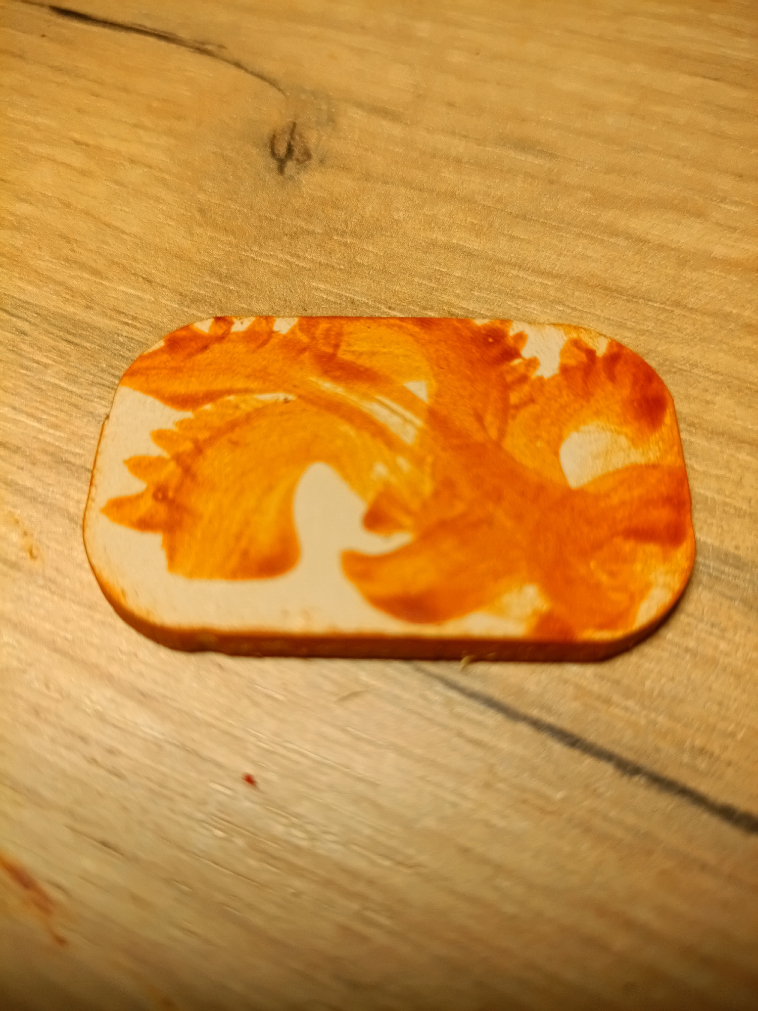 A close-up, eye-level shot shows a rectangular slice of white bread with rounded corners, placed on a light brown wooden surface. The bread has an orange, somewhat abstract pattern spread across its top surface, resembling smudges or a deliberate design. The wooden surface appears to be a table or countertop with visible grain patterns and a dark line running diagonally across it. The lighting casts a warm glow on the scene, with some shadows visible. The image is cropped to focus on the bread and a portion of the wooden surface. No other objects, people, or actions are depicted. The location in Irkutsk, Russia is not determinable from the image content.