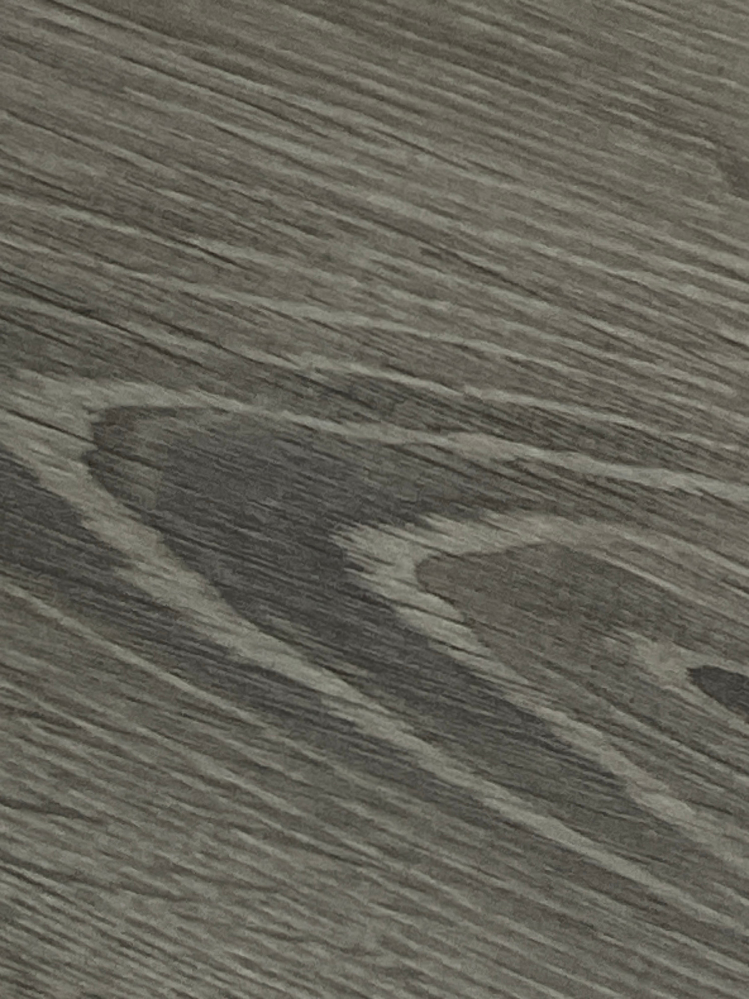 The image is a close-up, top-down view of a grey wood-grain textured surface. The grain runs diagonally from the bottom left to the top right, with darker lines forming a swirling, organic pattern in the center. The texture appears matte and possibly laminate or painted wood. There are no people, objects, actions, or interactions visible in the image. The location in Omsk, Russia, is not discernible from the image itself; it is a generic surface texture.