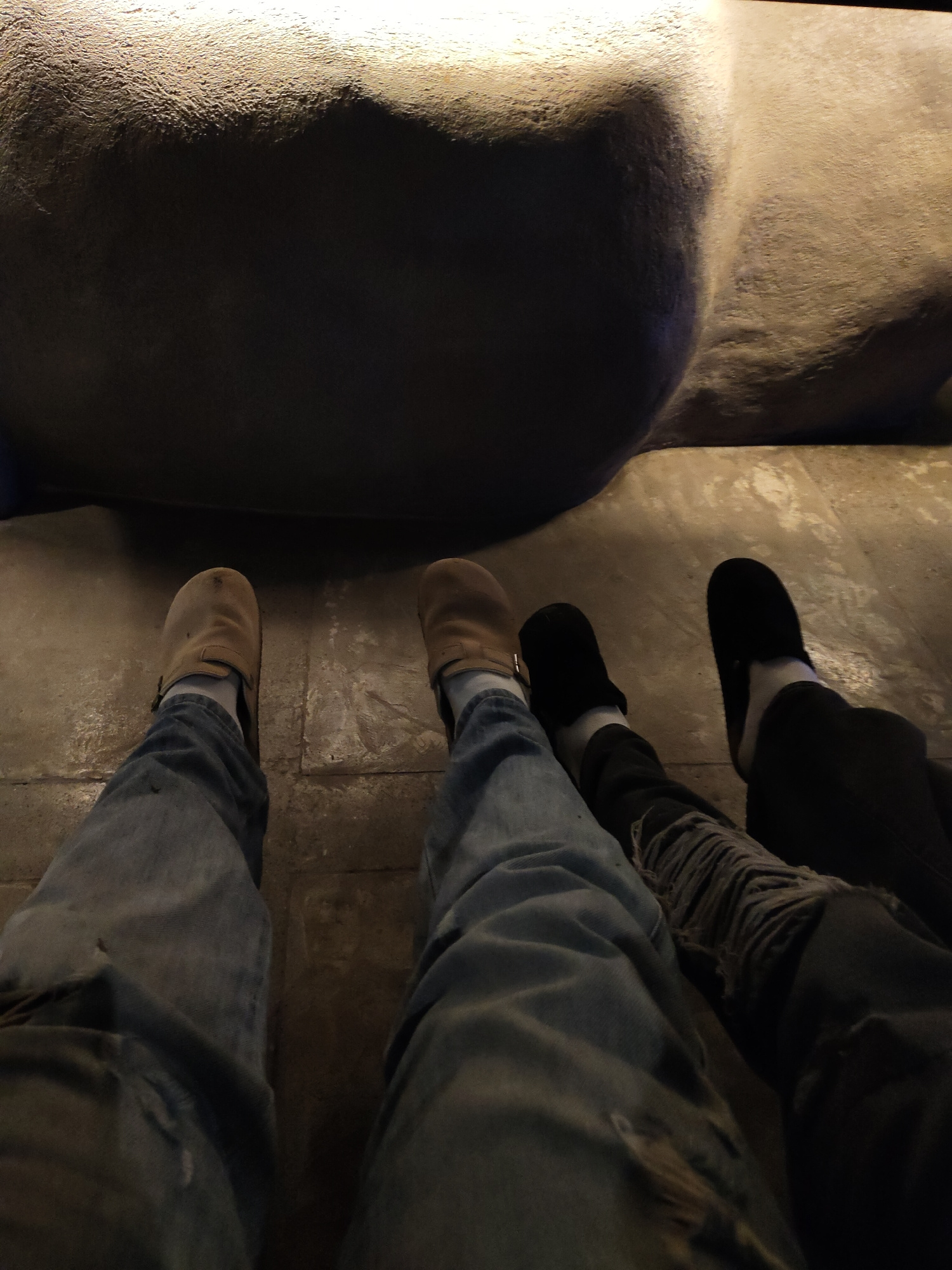 The image shows a low-angle perspective of two pairs of legs and feet, resting on a textured concrete floor. The left subject is wearing light blue jeans with rips and tears, and light brown clogs over white socks. The right subject is wearing dark pants, possibly denim with visible rips, and black slip-on shoes over white socks. Both subjects appear to be seated or lounging, facing forward or slightly away from the camera. In the background, a large, dimly lit, rough-textured object, possibly a rock formation or sculpture, dominates the upper portion of the frame, with the lighting highlighting its uneven surface. The environment appears to be indoors or in a shaded outdoor area, characterized by the concrete flooring and the artificial lighting. There is no specific indication of the location being in Davao City, Philippines, as the scene depicts common urban or recreational elements.