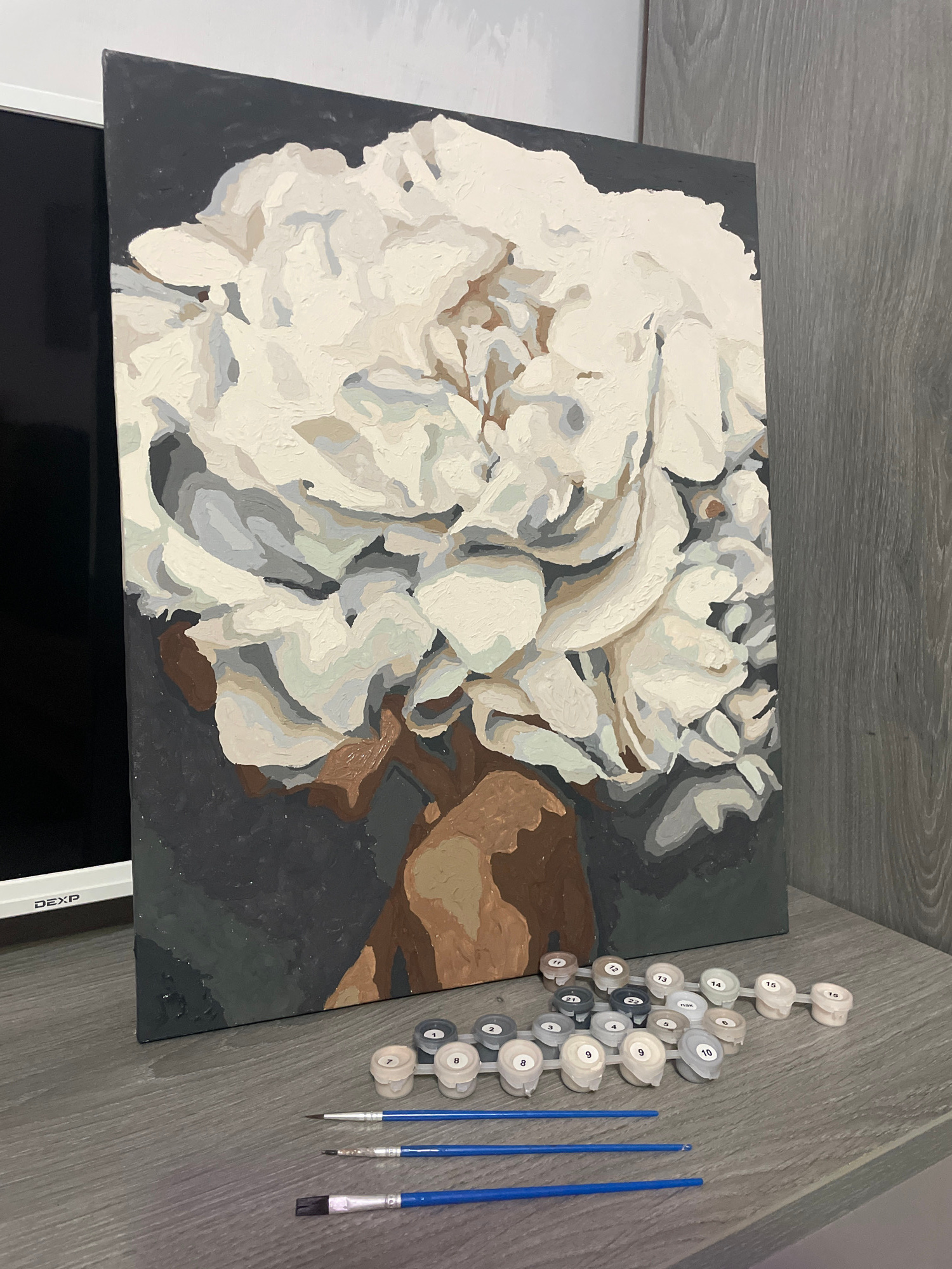 The image shows a paint-by-numbers kit, with the canvas featuring a partially completed painting of a white peony. The painting is rendered in shades of white, gray, and brown, with a dark gray background. To the right of the canvas are several small containers of numbered paint, a palette of numbered paint pots, and three brushes of varying sizes. The arrangement suggests that the painting is in progress. The canvas rests on a dark surface, possibly a table or easel, and is set against a background of gray wooden planks. This suggests an indoor setting. There is no visible text on the canvas itself, but one of the paint pots has a label that reads "TISSUE". The overall mood is one of creative activity, with the tools of the craft laid out alongside the emerging artwork.