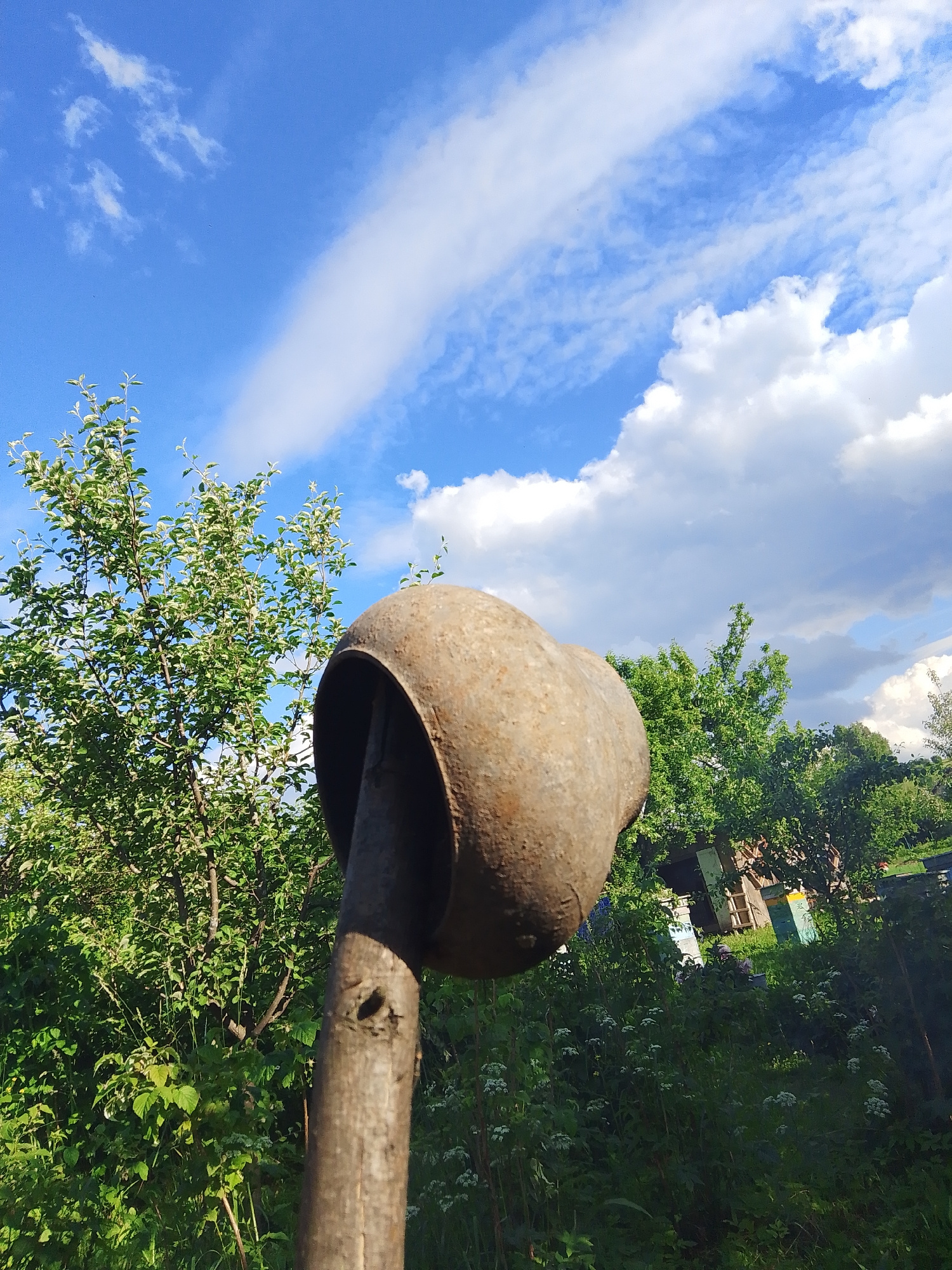 A close-up, low-angle shot captures a weathered, circular metal object attached to a wooden pole, dominating the foreground. The metal object appears to be rusty and hollow, resembling the top of a funnel or scoop. Behind it, a lush green tree with numerous leaves stands tall against a bright blue sky filled with dramatic white clouds. In the background, dense foliage and hints of structures, possibly bee hives, are visible, suggesting a rural or garden setting. The lighting indicates it is daytime, with sunlight casting shadows. The image is devoid of any human presence or activity.
