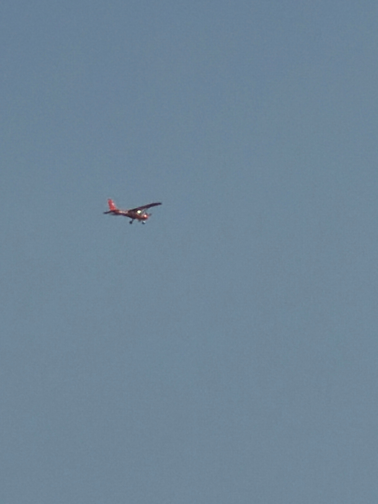 A small, red, single-engine airplane is captured in flight against a clear, pale blue sky. The aircraft is positioned in the upper left quadrant of the image, with its nose pointed slightly downwards. Its wings are spread horizontally, and its propeller is visible. The airplane appears to be a propeller-driven, fixed-wing aircraft. The sky is devoid of any clouds or other visible objects, suggesting an open-air environment. There is no information in the image to indicate a specific location in the city of Krasnoyarsk, Russia, or any other geographical context.