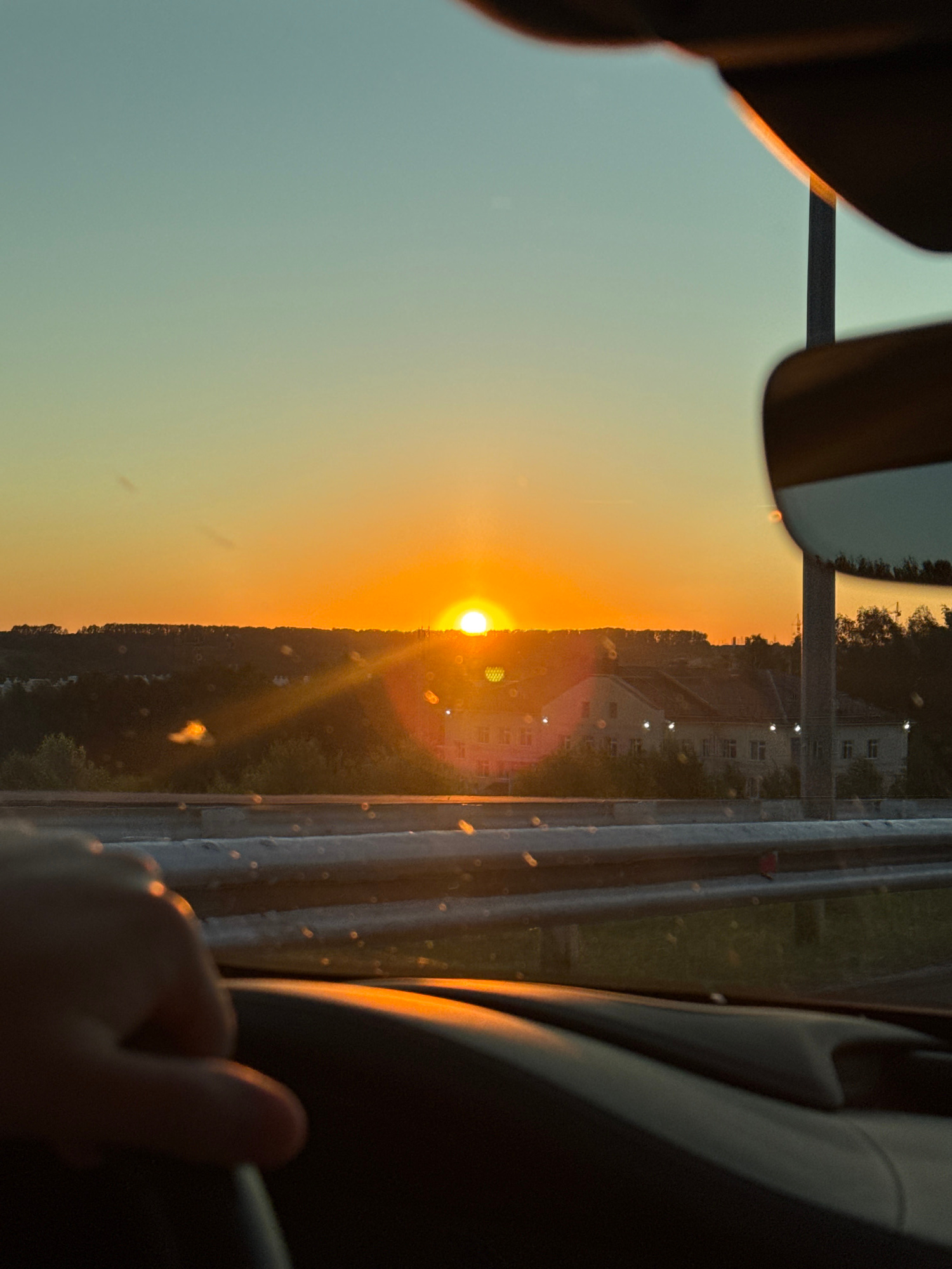 A vertical, eye-level, slightly high-angle shot captures a sunset viewed from inside a vehicle. The sun, a bright, orange orb, is centered on the horizon, casting a warm glow and a lens flare across the scene. The sky transitions from a pale blue at the top to a vibrant orange near the horizon.
Below the sky is a hilly landscape dotted with trees and houses. The houses appear to be of a traditional architectural style, with light-colored facades and red roofs, clustered together. A line of trees forms a dark silhouette along the crest of the hills.
In the foreground, a hand is visible on the steering wheel, indicating the viewer is the driver. The interior of the car is dark, with the dashboard and a portion of the windshield frame visible. A side-view mirror on the right reflects a portion of the sky and a light pole. A metal guardrail runs along the edge of what appears to be a highway or elevated road. The overall impression is of a serene journey during golden hour, with the natural beauty of the sunset dominating the view.