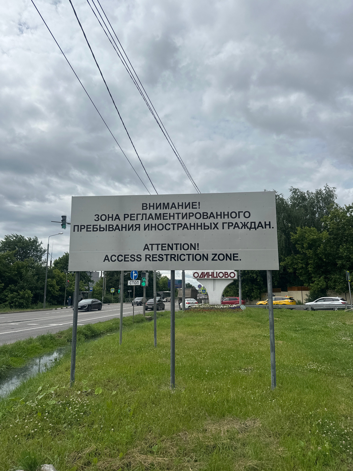 An outdoor, medium-shot image shows a large, white, rectangular sign on two metal poles in the foreground, with a cloudy sky and greenery in the background. The sign has text in both Russian and English. The Russian text reads: "ВНИМАНИЕ! ЗОНА РЕГЛАМЕНТИРОВАННОГО ПРЕБЫВАНИЯ ИНОСТРАННЫХ ГРАЖДАН." The English text reads: "ATTENTION! ACCESS RESTRICTION ZONE." Below the main sign, there is another sign that says "ОДИНЦОВО".
To the left of the sign, there is a traffic light that is green. There are also power lines strung across the top of the frame. A road is visible in the middle ground with several cars driving on it, including a black sedan, a red car, a yellow taxi, and a white sedan. There is a grassy median between the road and the sign, with some small white flowers scattered throughout. A ditch with murky water is visible to the left of the sign. Trees and buildings are in the background. There are no people clearly visible in the image.