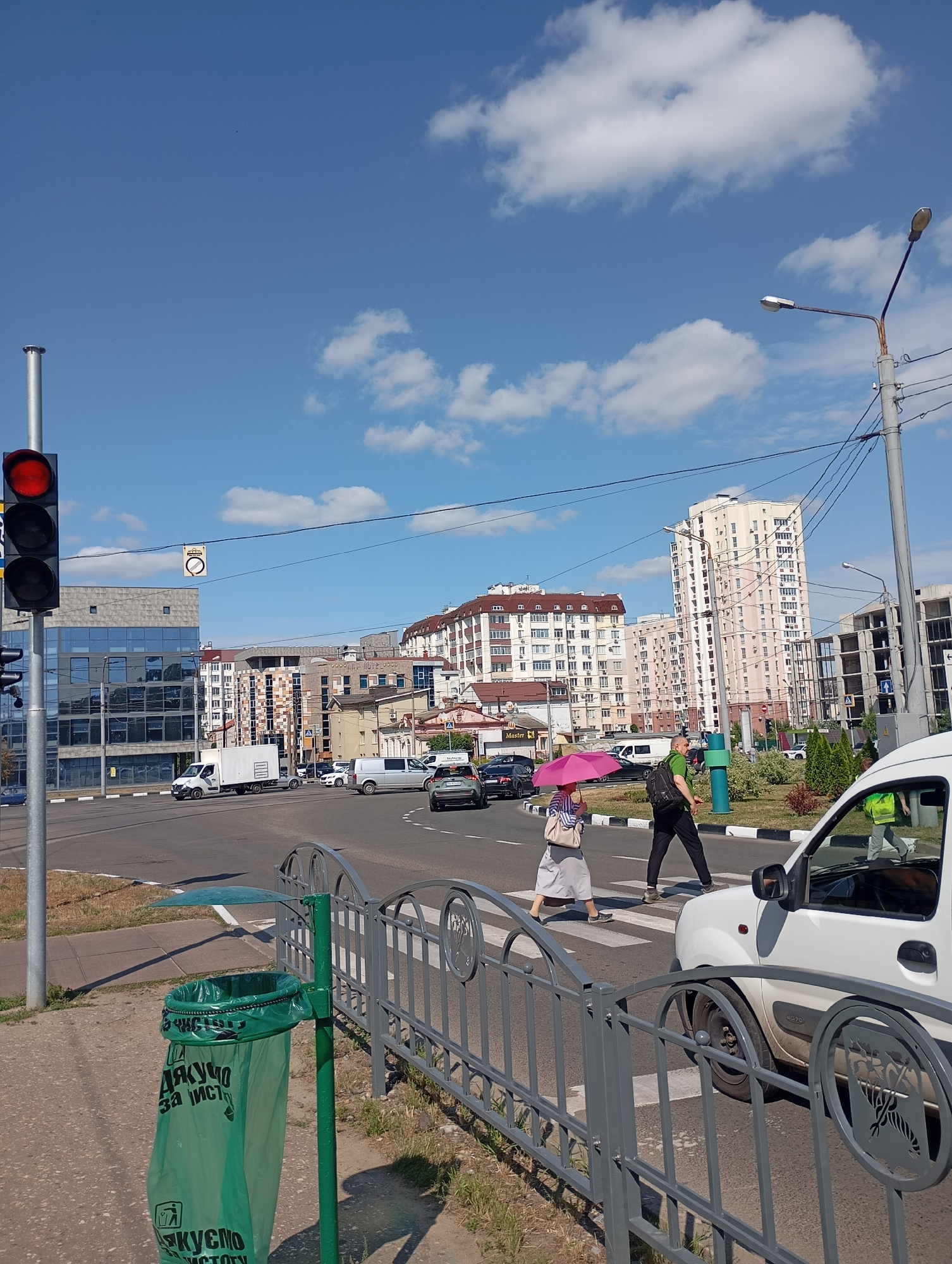 The image depicts a street scene in Kharkiv, Ukraine, under a clear blue sky with scattered clouds. In the foreground, a green trash bin with Cyrillic text reading "ДЯКУЮ ЗА ЧИСТОТУ" (Thank you for cleanliness) is visible on a sidewalk, next to a gray metal fence. Across the street, a pedestrian crossing is marked by white stripes. A woman with a pink umbrella and a man with a backpack are crossing the street. A white car is stopped on the right side of the frame, partially obscuring the pedestrian crossing. In the background, several buildings are visible, including a modern multi-story residential building with a light-colored facade and balconies, and other apartment buildings. A traffic light, showing red, is positioned on the left side of the image, along with a street lamp. Vehicles, including a white van and several cars, are present on the road, indicating traffic flow. The overall impression is of an urban environment on a sunny day.
