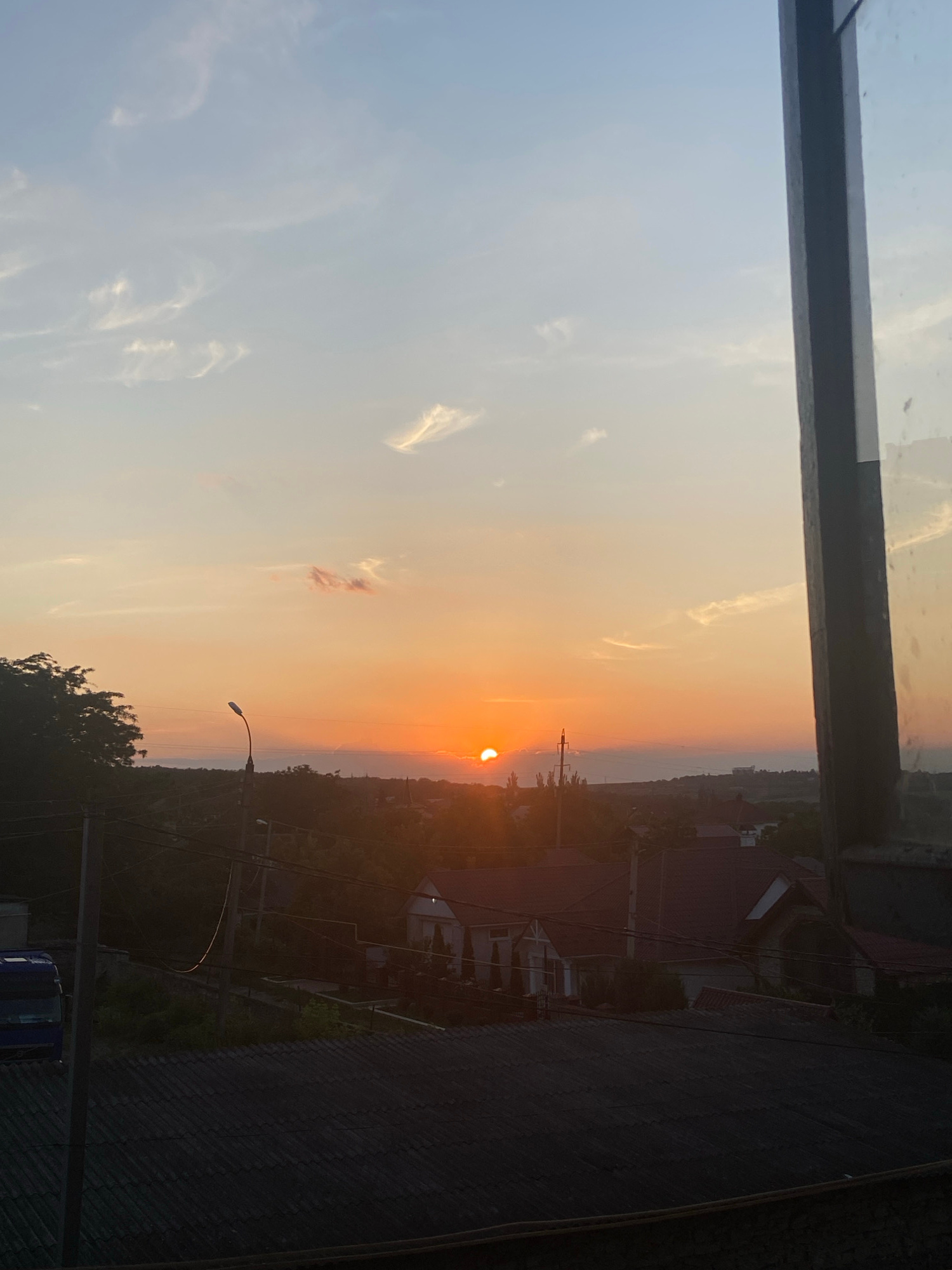 A wide-angle, over-the-shoulder shot captures a sunset viewed through a window. The sky is a gradient of soft blues and oranges, with wisps of clouds scattered across it. The sun, a bright orange orb, is low on the horizon, casting a warm glow over the landscape. In the foreground, the dark, textured surface of a rooftop is visible, partially obscuring the view. Beyond the rooftop, houses with red-tiled roofs are nestled amongst lush green trees. A lone streetlamp stands tall, its curved neck reaching towards the sky. Electrical wires crisscross the scene, adding a layer of urban infrastructure to the natural beauty of the sunset. A blue vehicle is parked on the left side of the frame, partially visible behind a utility pole.