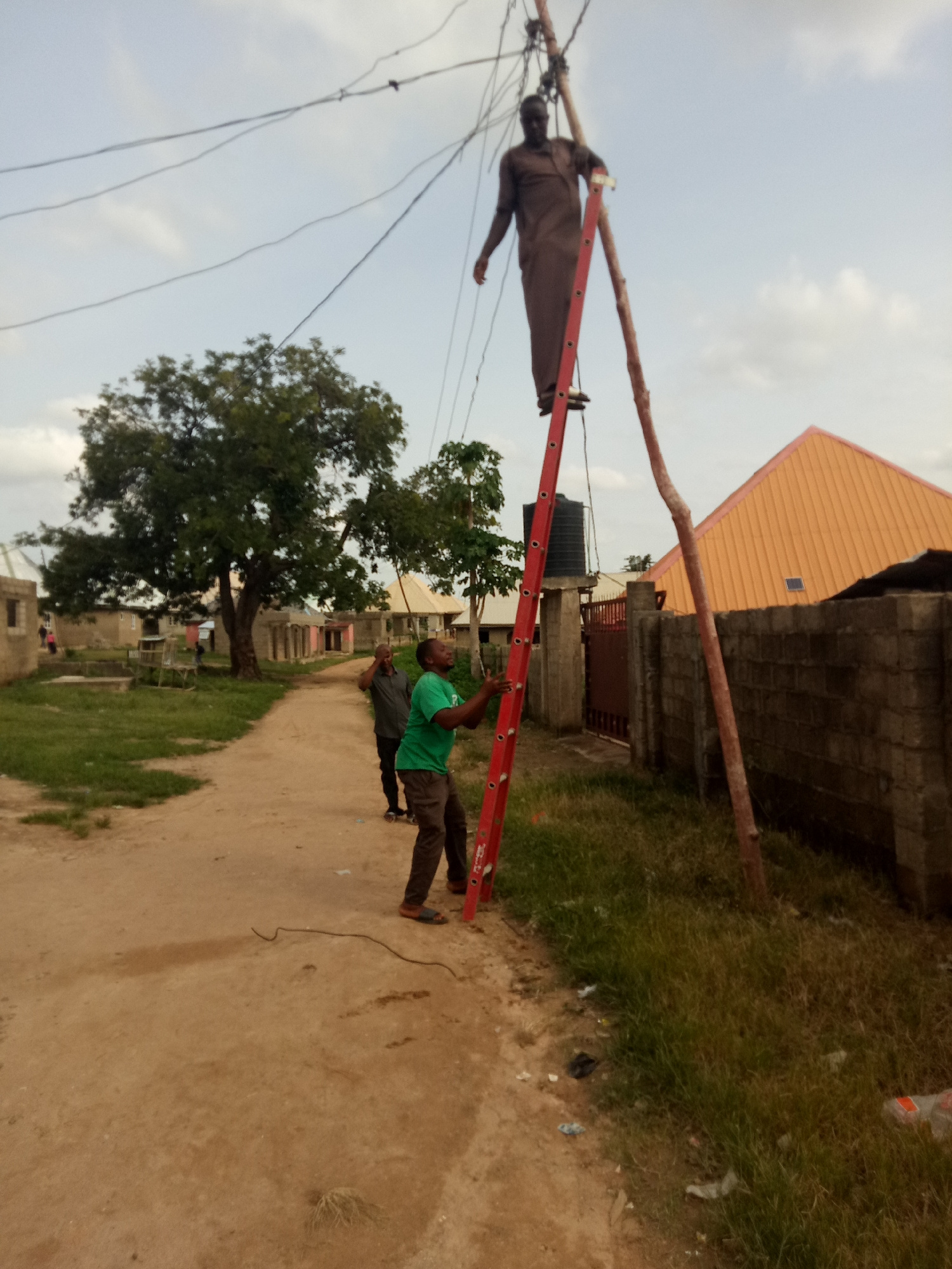 In an outdoor, semi-urban environment, three individuals are engaged in what appears to be electrical work. A man, dressed in a brown traditional garment, is positioned precariously on a tall red ladder, reaching towards overhead wires. Another man, wearing a green t-shirt and dark trousers, stands on the ground, also holding the ladder, likely for stability. A third man, in a grey shirt and dark trousers, stands behind the man in green, looking up at the scene. Overhead, a network of electrical wires is visible against a cloudy sky. The ground is a dirt path, with some sparse grass growing along the side. In the background, there are buildings with corrugated roofs, a stone wall, and a large tree. The overall scene suggests a communal area where essential services are being maintained.