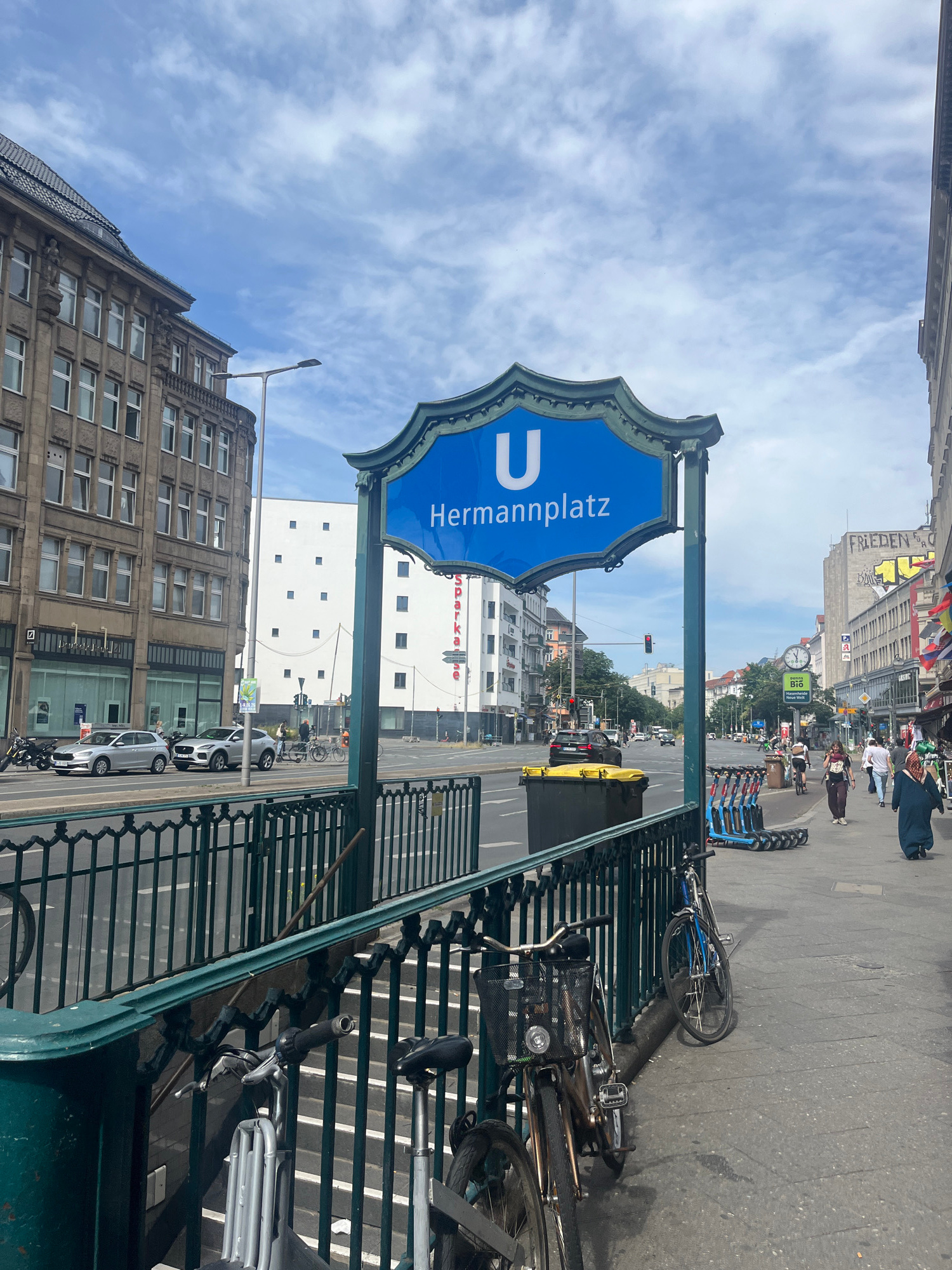 The image captures a street scene in Berlin, Germany, specifically at the U-Bahn (subway) station "Hermannplatz." A prominent blue sign with white lettering designates the station entrance, featuring a stylized "U" for Untergrundbahn. In the foreground, a dark green metal railing separates the subway entrance from the sidewalk. Several bicycles are parked near the railing, and a trash bin is visible.
The sidewalk to the right of the railing is populated with pedestrians. Some are walking, while others appear to be waiting or gathering. A row of electric scooters is parked along the sidewalk. Buildings line the street on both sides. On the left, a multi-story building with a stone facade displays numerous windows. Across the street, more buildings are visible, including a modern white structure with square windows and older buildings with shops at ground level.
The street itself is active with vehicular traffic, including cars and motorcycles. Traffic signals are visible, indicating an intersection. The sky above is partly cloudy, with a mix of blue sky and white clouds. The overall scene depicts a bustling urban environment with public transportation, pedestrian activity, and commercial establishments.