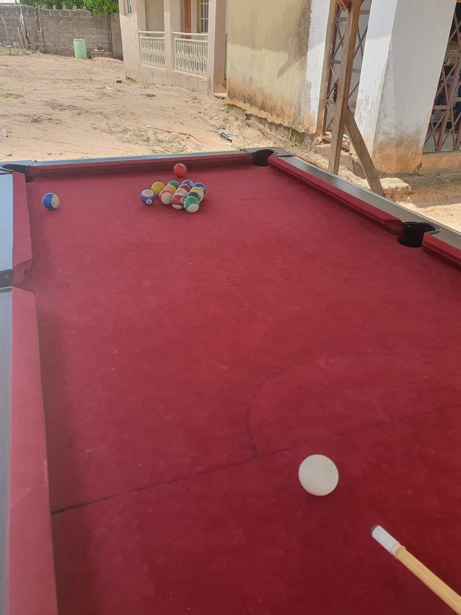 This image captures a pool table with several billiard balls and a cue stick, set against an outdoor backdrop. The pool table is covered in red felt, with some balls clustered together and others spread out. One white ball is positioned near the cue stick, suggesting an ongoing game or a paused moment of play.
The scene appears to be an outdoor area in Lokoja, Nigeria, as indicated by the surroundings. Behind the pool table, there is a building with multiple levels and balconies, constructed with concrete and possibly some metallic elements. The ground is a dirt or sandy surface, with scattered debris and some vegetation. The lighting suggests it is daytime, and the overall atmosphere seems casual and informal, likely a leisure setting.
Notable details include the arrangement of the billiard balls, some of which are numbered and colored according to standard pool games. The cue stick is white with a beige handle. The building's architecture, with its balconies and white railings, provides a glimpse into the local style. The presence of dirt and some untidiness in the foreground suggests an outdoor environment that might not be meticulously maintained, which is common in many outdoor recreational areas. There is no visible text in the image.