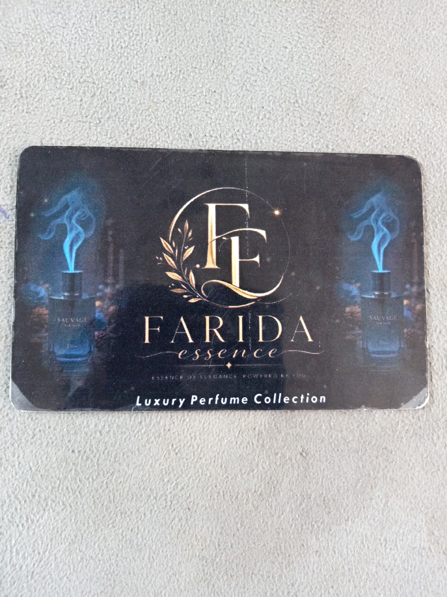 The image displays a black gift card with a glossy finish, featuring a brand logo and product imagery. The brand logo consists of stylized golden letters "FF" enclosed in a circular laurel wreath. Below the logo, the brand name "FARIDA" is printed in large, serif font, followed by "essence" in a smaller, cursive font. Further down, in a smaller, distressed font, is the text "ESSENCE OF ELEGANCE. POWERED BY YOU." At the bottom of the card, in white capital letters, is the text "Luxury Perfume Collection".
On either side of the brand logo are images of perfume bottles emitting blue smoke. The bottle on the left is labeled "SAUVAGE". Both bottles are dark glass and appear to be luxury fragrance products. The background of the card is dark and speckled, suggesting a night-time or ethereal theme.
The card is placed on a textured, light gray surface, possibly concrete or a similar material. The lighting suggests an indoor environment with overhead light casting faint shadows. The image is taken from a slightly elevated angle, looking down at the card. There are no people or overt actions depicted in the image. The setting is an unspecified indoor location, with no discernible urban or environmental context that would place it within a specific city or area.