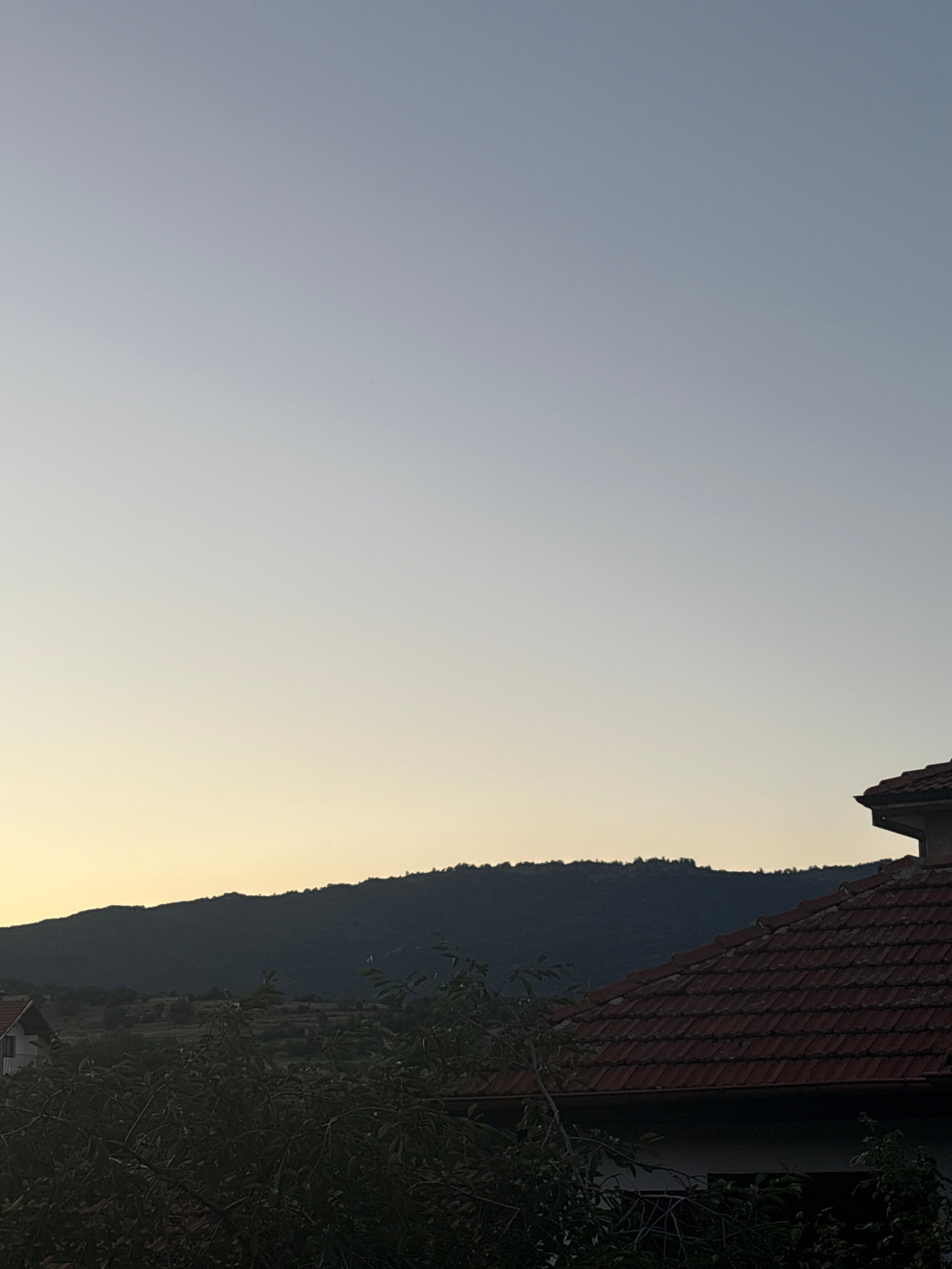 The image depicts a dusky landscape with a clear, gradient sky transitioning from light blue at the top to a pale yellow near the horizon. A dark, silhouetted mountain range with a textured treeline forms the middle ground. In the foreground, the terracotta tiled roof of a house is visible on the right, with dark green foliage obscuring the lower portion of the scene. A white building with a red roof is partially visible on the left. The overall impression is a serene, rural view, possibly from a vantage point overlooking the town of Strelcha, Bulgaria, at dusk.