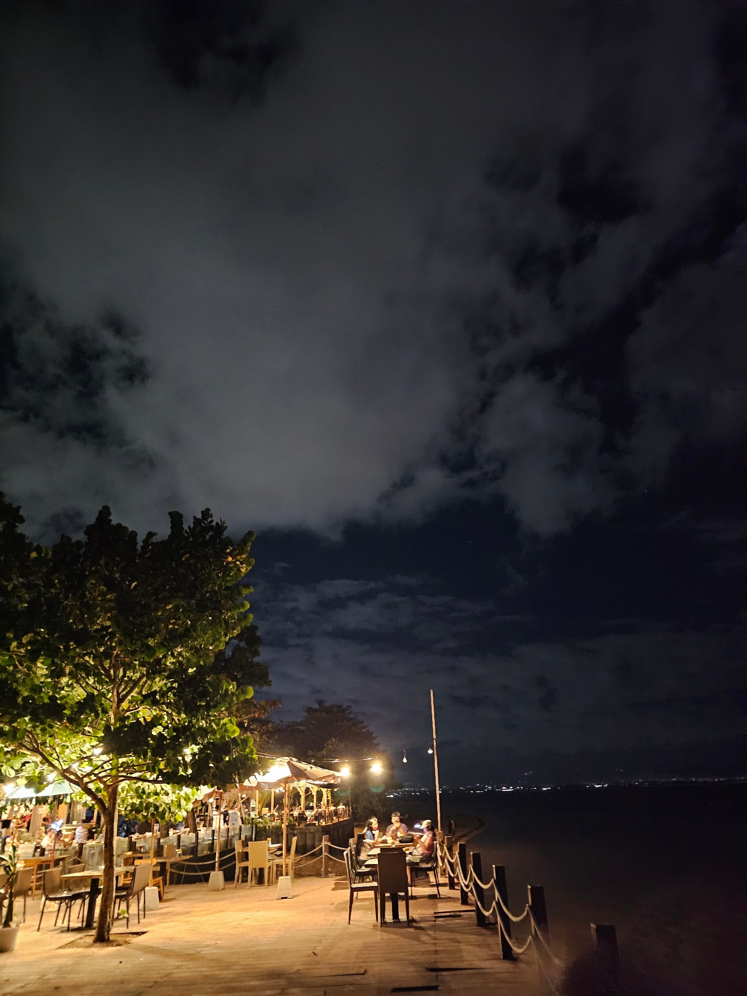 The image depicts a nighttime scene at a beachfront restaurant in Denpasar, Indonesia. A wooden boardwalk runs along the edge of the water, lined with tables and chairs illuminated by warm lighting. A large tree with lights strung through its branches stands prominently on the left side of the boardwalk. Several people are seated at tables, appearing to be dining and socializing. Umbrellas provide shade over some of the seating areas, and string lights are visible in the background, adding to the ambiance. The ocean is visible to the right, and a distant city skyline with lights can be seen across the water. The sky is dark and cloudy.