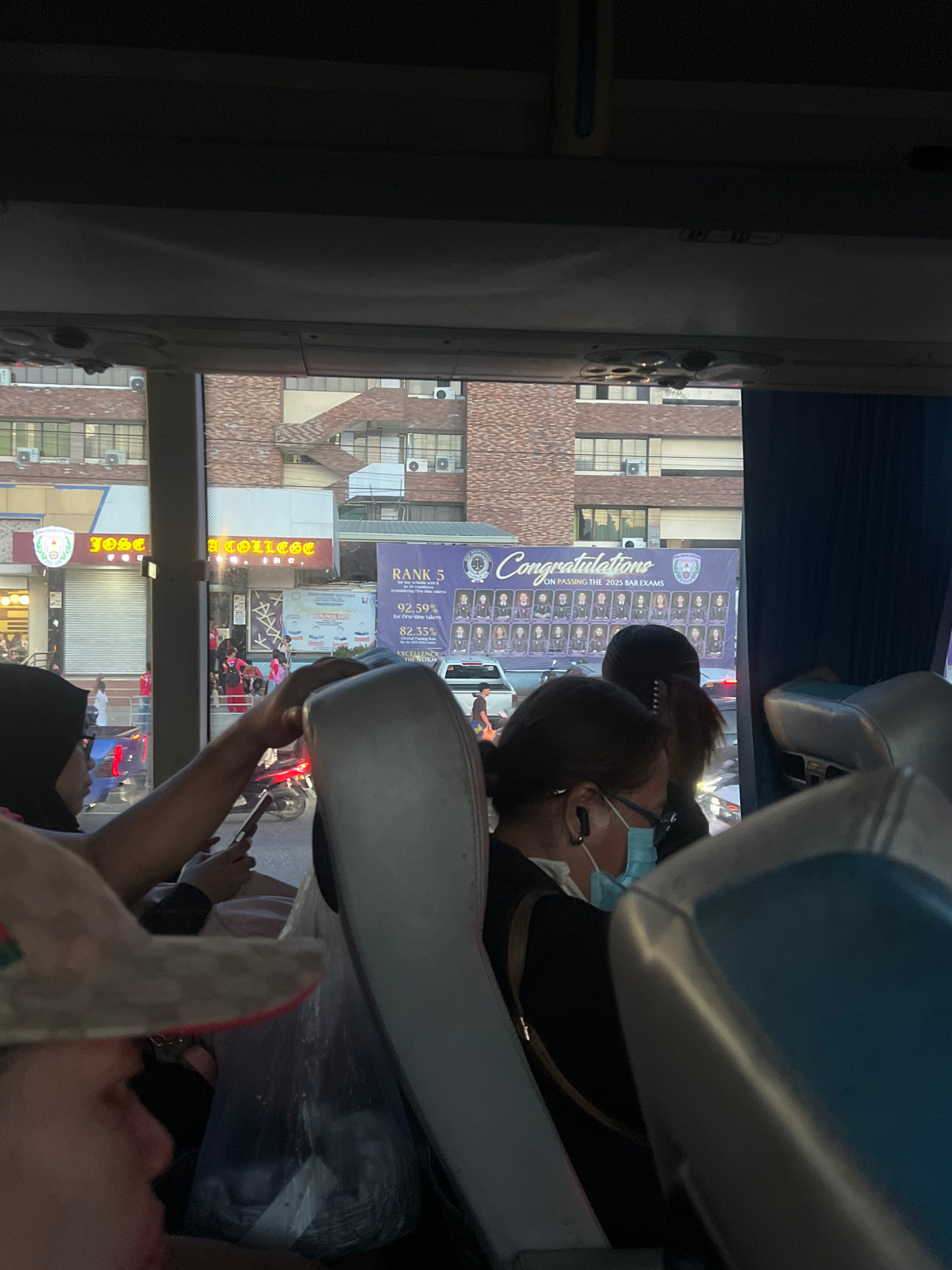 The image shows a view from inside a vehicle, possibly a bus, looking out a window at a street scene in Davao City, Philippines. In the foreground, the heads and shoulders of several passengers are visible, some wearing masks. One passenger is holding a mobile phone. Outside the window, a building with the sign "JOSE R. URBINA COLLEGE, INC." is prominent. Adjacent to it is a large banner congratulating graduates on passing the 2025 Bar Exams, with numerous individual photos of the graduates. The street appears busy with traffic, including cars and motorcycles. The architecture suggests an urban environment.