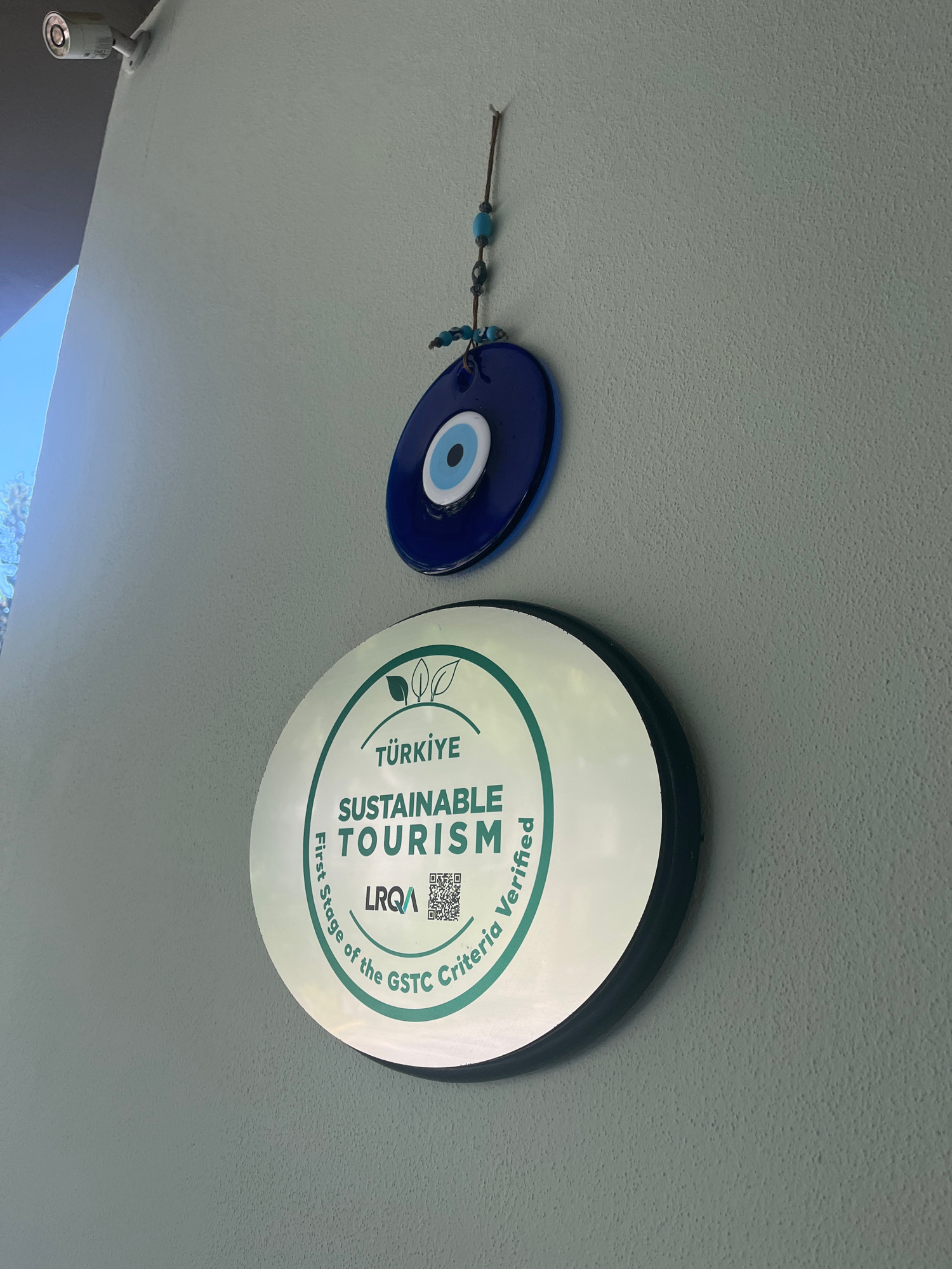 This image shows a security camera mounted on a textured wall in Alanya, Turkey. Below the camera, a blue evil eye charm with concentric circles of white, light blue, and dark blue hangs from a string. Further down the wall is a circular, green-and-white plaque. The plaque has the words "TÜRKIYE SUSTAINABLE TOURISM" printed in green text, along with the LRQA logo and a QR code. The words "First Stage of the GSTC Criteria Verified" are printed in a circular pattern around the edge of the plaque. The wall is painted a light gray color, and the background shows a bright blue sky on the left side of the frame.
