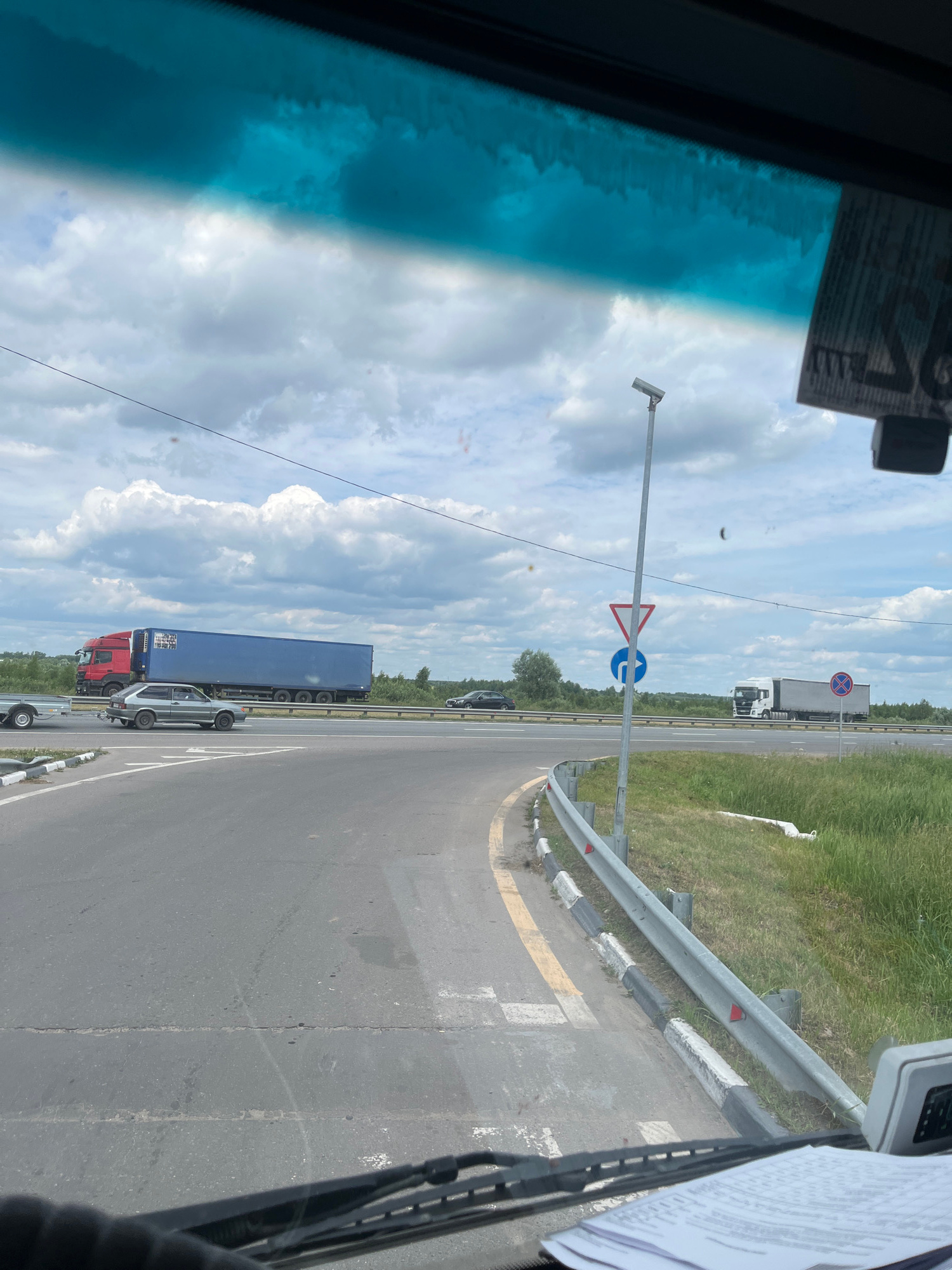 The image is taken from inside a vehicle, looking forward through the windshield. The scene is a road intersection in Старое Бобренево, Russia, under a cloudy sky. On the left, a grey passenger car is seen driving on a paved road. In the middle ground, a large red truck with a blue trailer is driving on a highway. Further to the right, another white truck with a grey trailer is also on the highway. Road signs are visible, including a give way sign, a blue directional arrow sign, and a red and white circular prohibition sign. Guardrails line the side of the road, and tall green grass and trees are visible in the background. The interior of the vehicle shows a dashboard with papers and a piece of equipment.