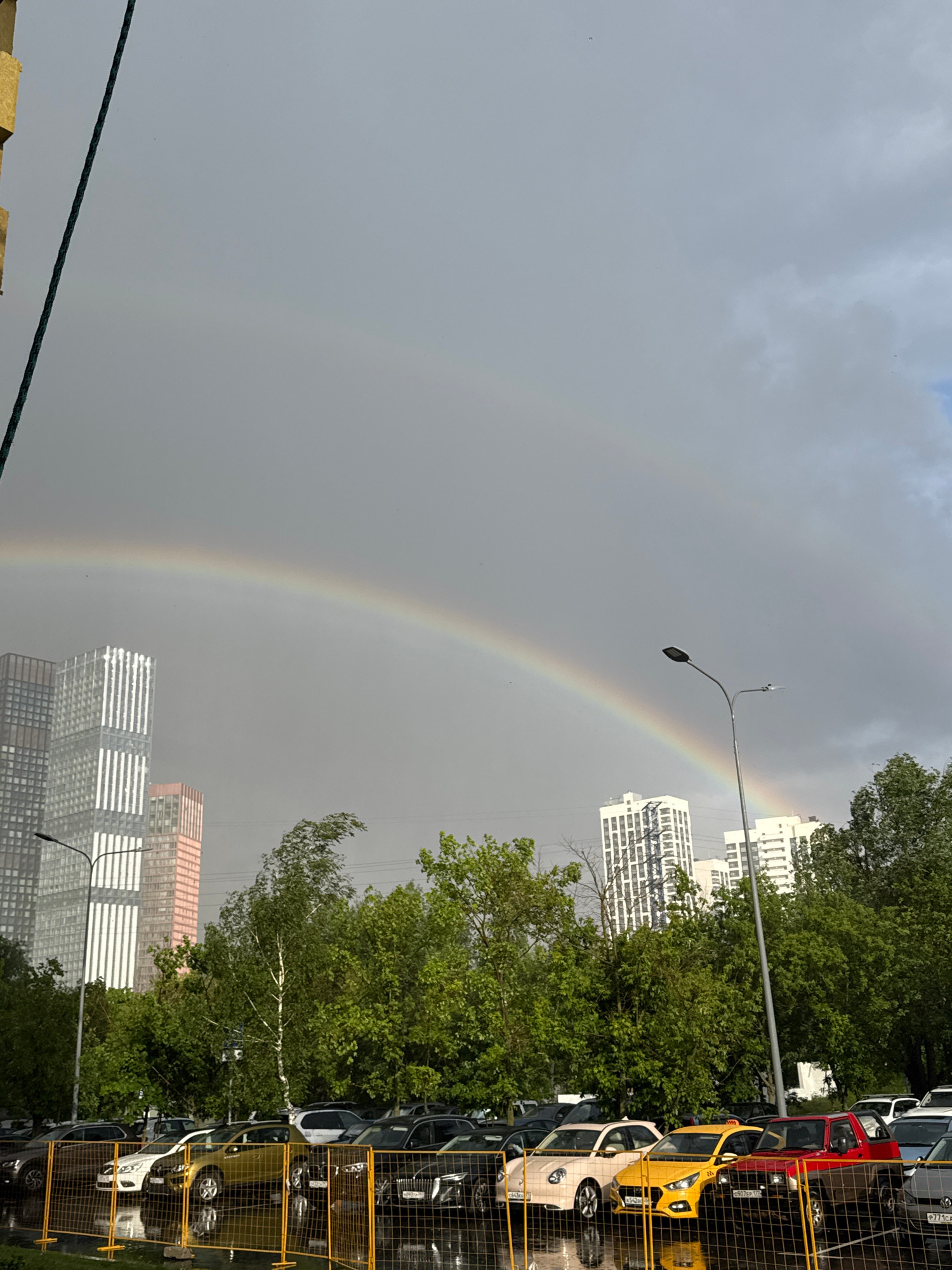 The image shows a double rainbow arching across a gray, overcast sky. The rainbow is partially obscured by trees and buildings. On the right side of the image, a row of parked cars is visible behind a yellow mesh fence. The cars are of various colors, including yellow, red, and white. Some of the cars have visible license plates, but the text is illegible. In the background, several modern high-rise buildings can be seen, some with a pattern of horizontal lines on their facades. A street lamp with a single light fixture is also present in the scene. The overall impression is of an urban environment, possibly during or after a rain shower, given the overcast sky and the presence of a rainbow. No people are visible in the image. The time of day is uncertain due to the overcast conditions, but the presence of a rainbow suggests sunlight is breaking through the clouds.