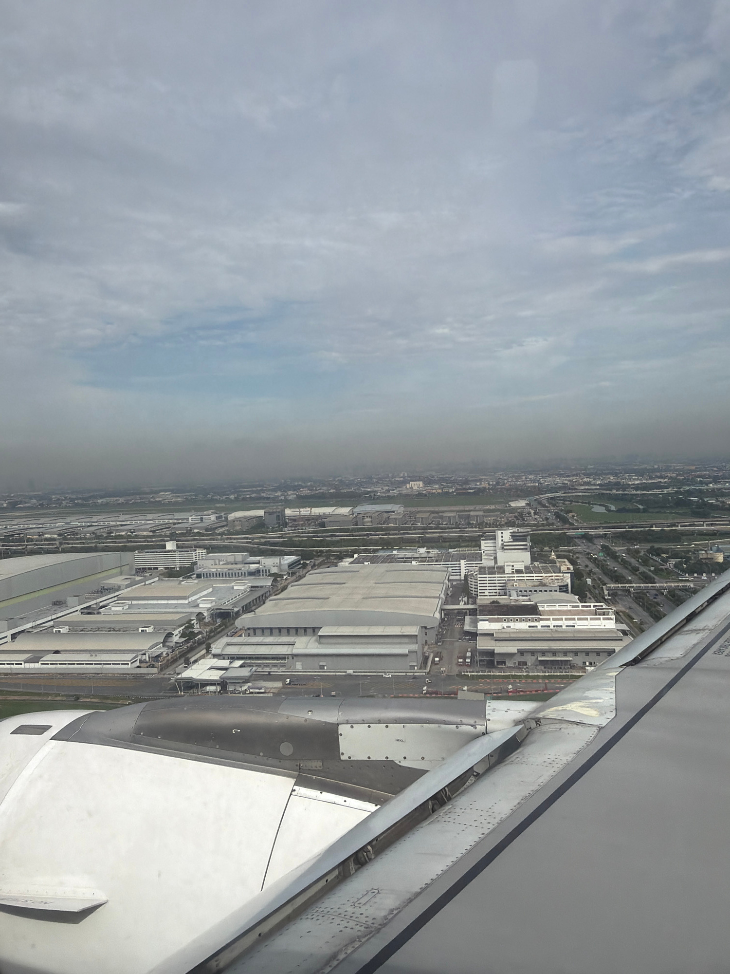 An aerial view captures a sprawling industrial complex under a hazy sky, likely taken from an airplane. The foreground shows a portion of an airplane's wing and engine. The industrial area features numerous large, flat-roofed buildings with metallic or light-colored exteriors, arranged in a grid-like pattern. Roads and transportation infrastructure, including overpasses and what appear to be train tracks, weave through the complex. In the distance, a city skyline is visible, partially obscured by atmospheric haze. The overall environment suggests a heavily developed urban or suburban industrial zone. The location is identified as Nong Prue Subdistrict, Thailand. There are no visible people or specific actions depicted in the image.