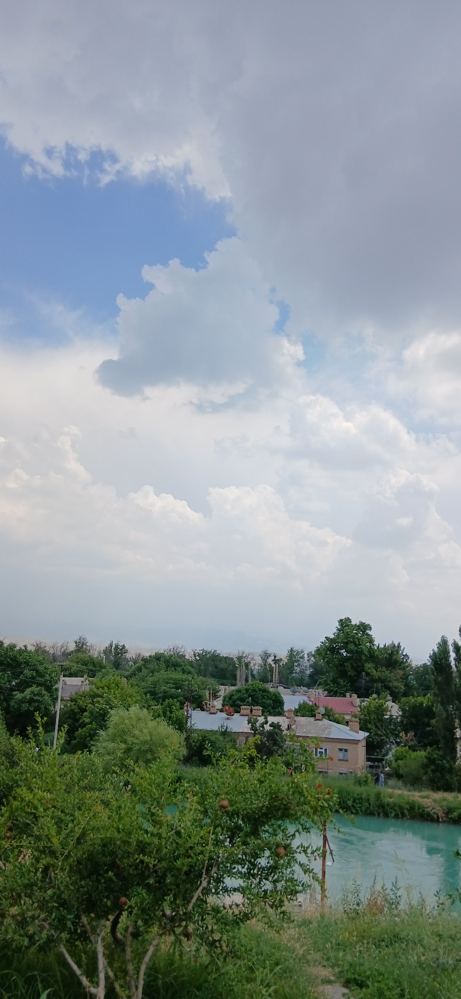 The image depicts a landscape view from an elevated position, overlooking a body of water and a residential area. The sky is filled with dramatic cumulus clouds, with patches of blue sky visible. In the foreground, lush green foliage and a tree with visible fruit, possibly pomegranates, dominate the lower left of the frame. A bright turquoise body of water lies to the right of the foliage. Beyond the water, rooftops of houses are visible, interspersed with trees. Further in the distance, a hazy outline of a cityscape or more distant terrain is visible under the cloud cover. The overall impression is of a serene, perhaps rural or suburban, environment on a cloudy day.